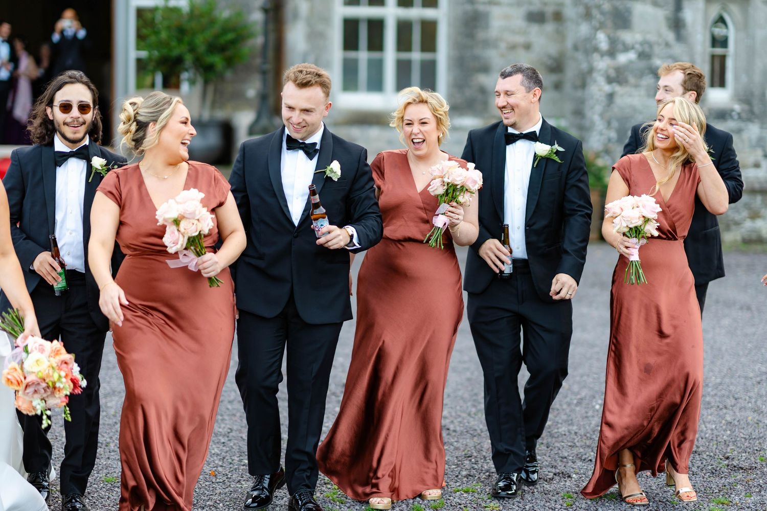 Wedding party in black tuxedos and rust-colored bridesmaid dresses walk and laugh together outdoors.