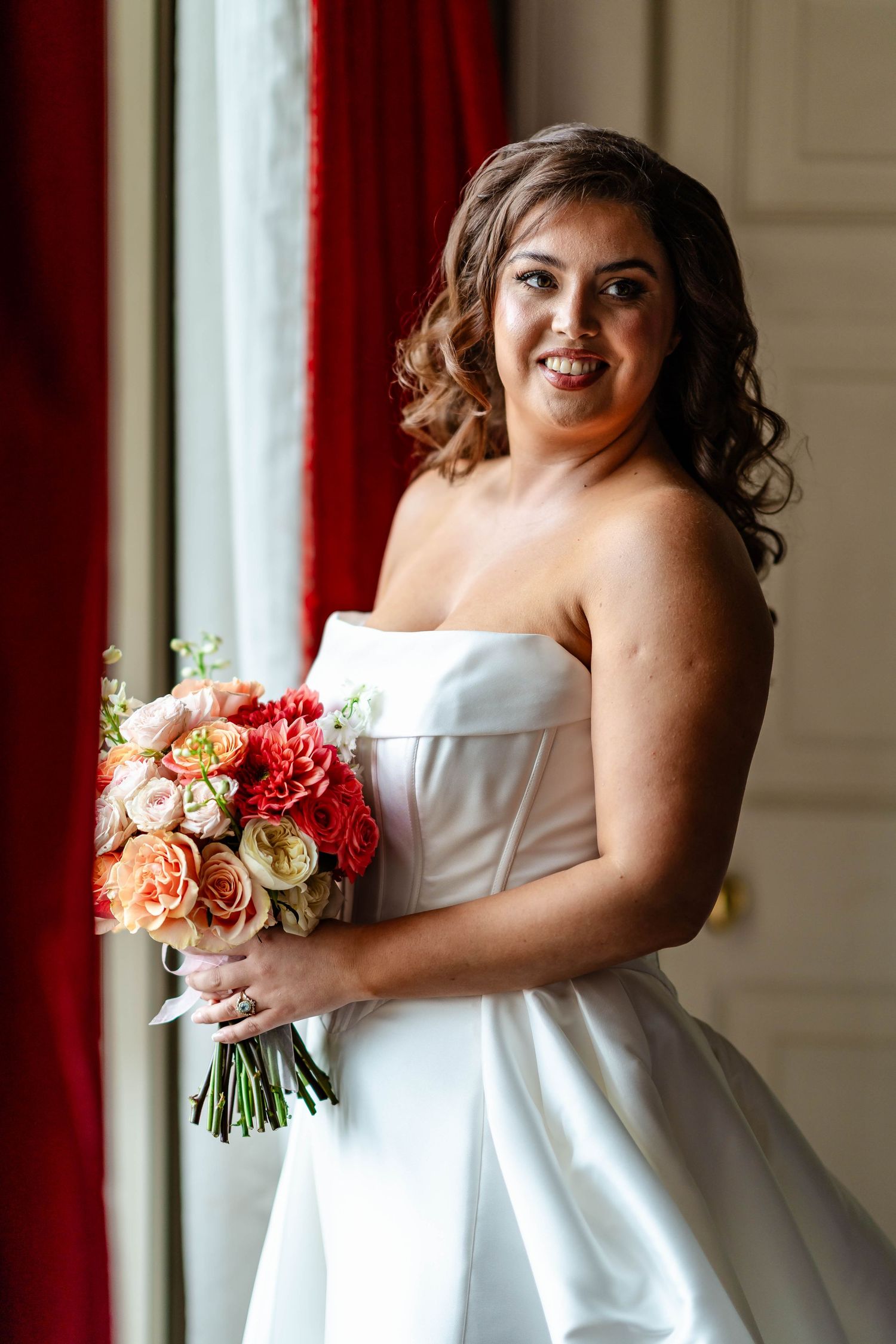 Bride in strapless white ballgown holds vibrant coral and peach bouquet while standing by window with red curtains.
