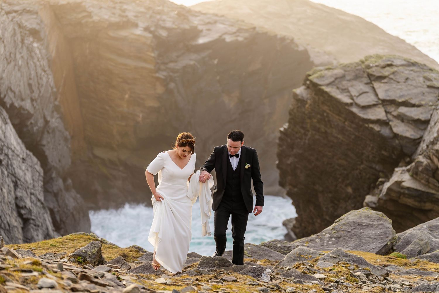 Newlyweds share an intimate moment on rocky cliffs overlooking turquoise ocean waters.