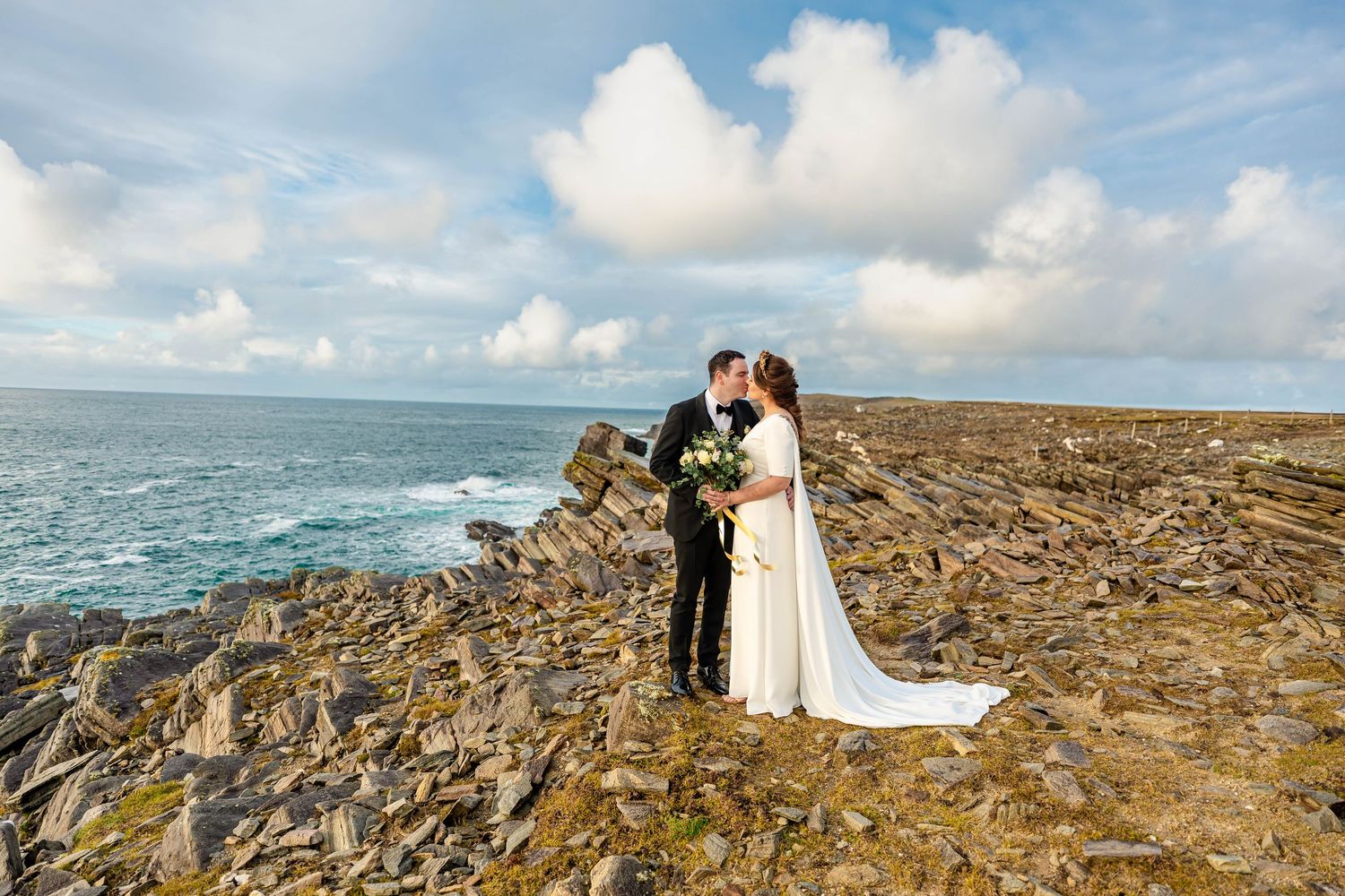 Romantic wedding moment on rocky ocean cliffs with waves crashing below.