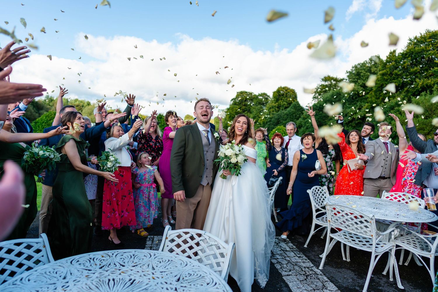 Wedding guests throw confetti during an outdoor celebration with white garden furniture.
