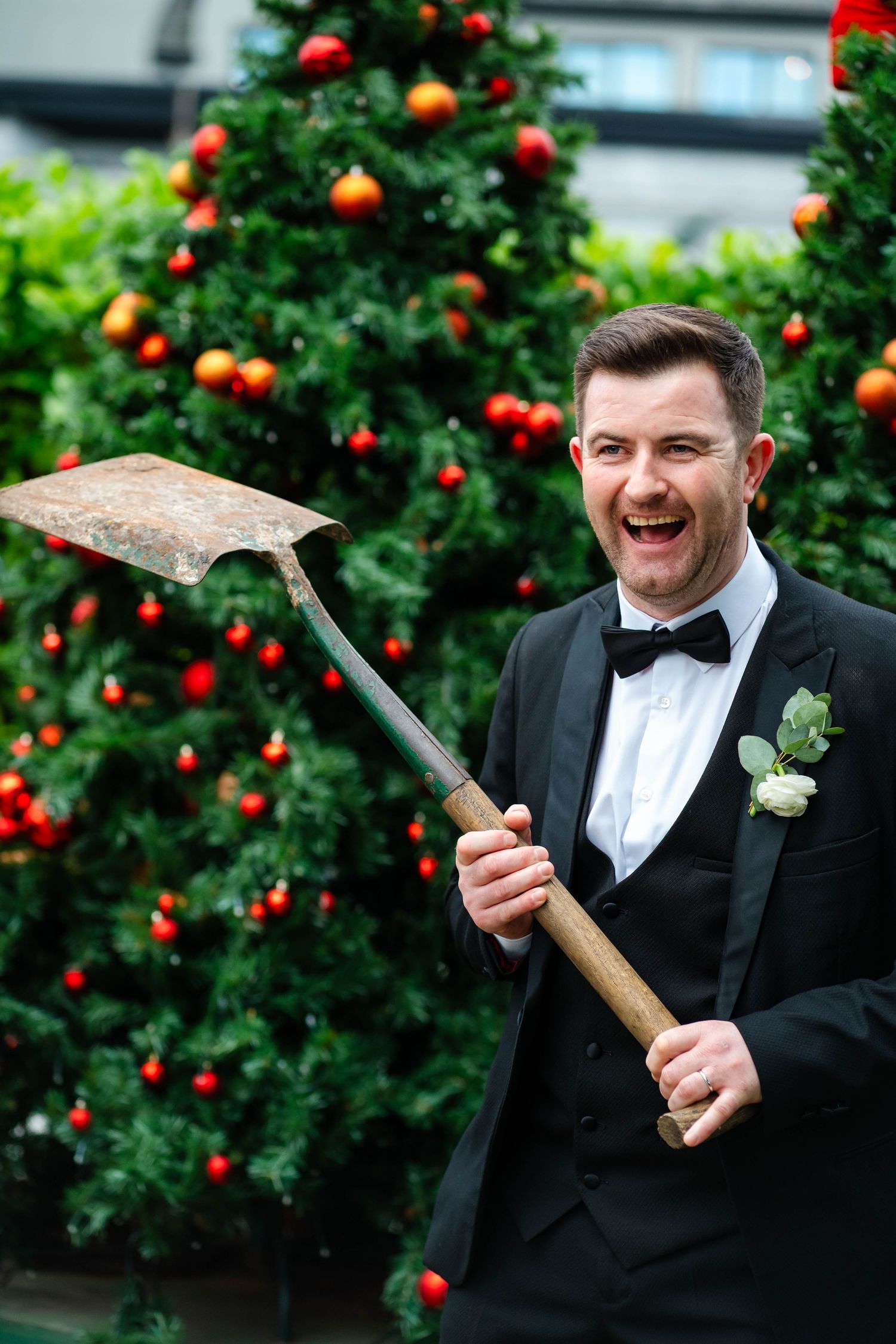 Groom in black tuxedo playfully poses with a shovel against a backdrop of red ornaments and greenery.