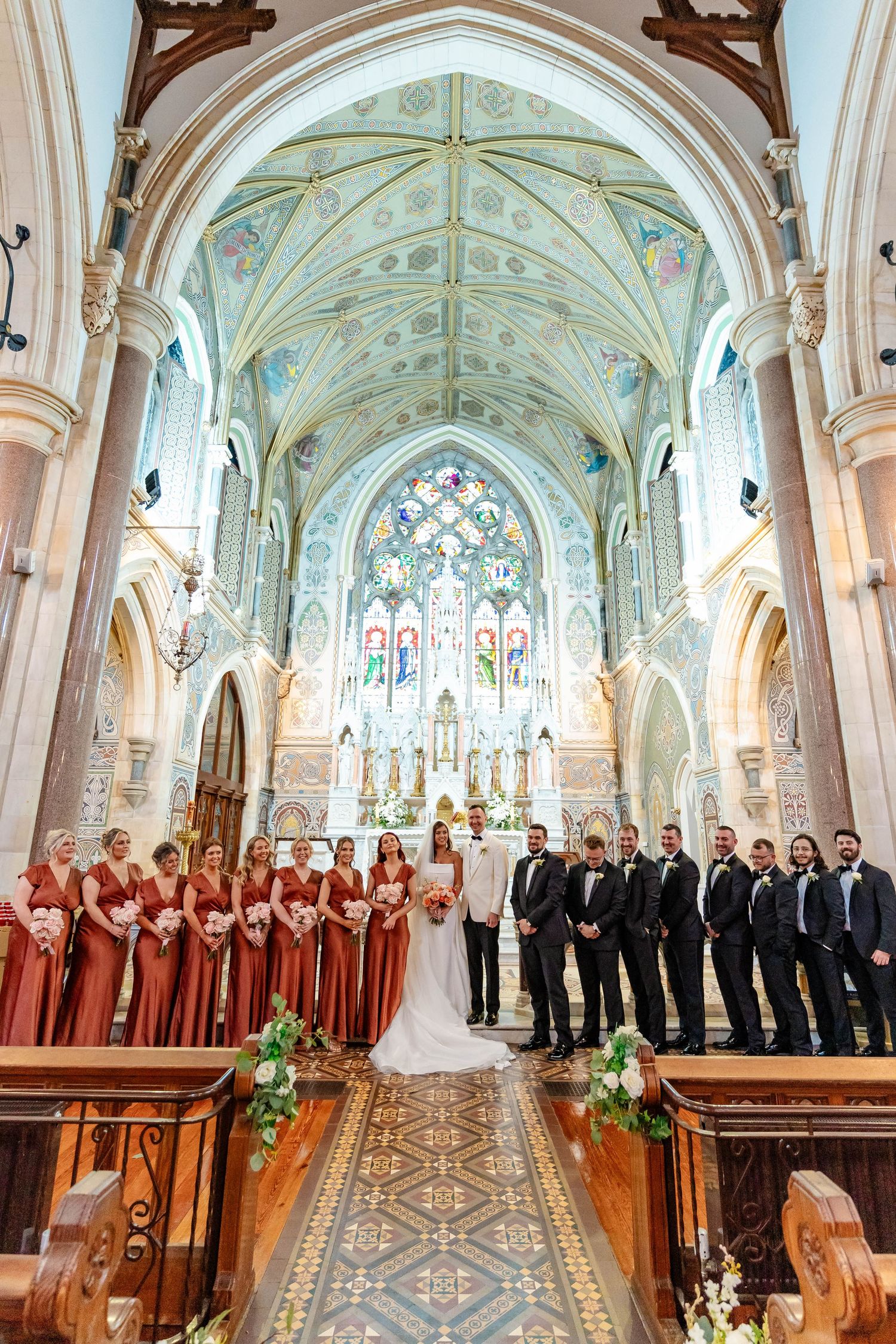 Wedding party stands in ornate Catholic church with vaulted ceilings, stained glass windows and decorative architectural details.