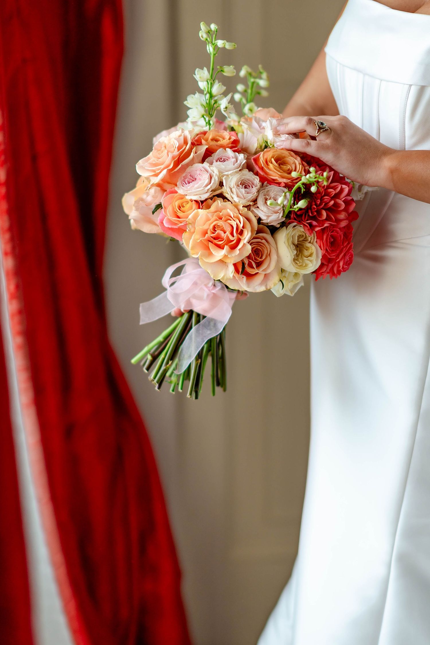 Elegant wedding bouquet with coral and peach roses against red curtain backdrop and white dress.
