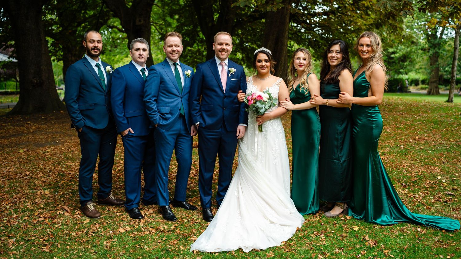 A wedding party gathers for photos on a tree-lined path with fallen leaves.