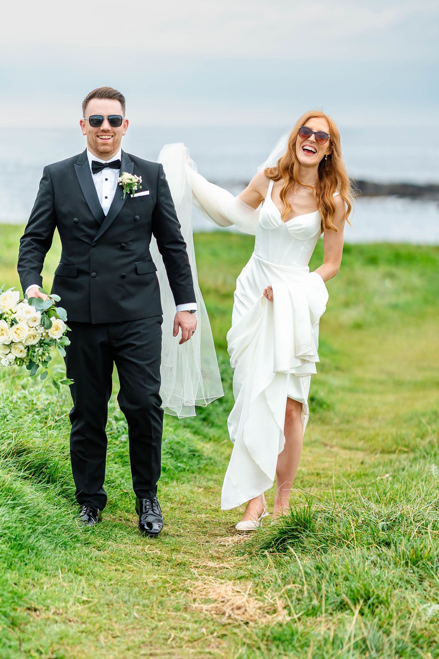A couple walks along a grassy coastal path in formal wedding attire with a bouquet of flowers.