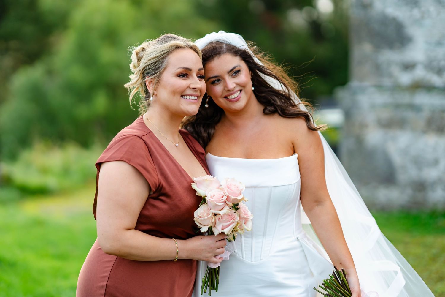 A bride in a white wedding gown and bridesmaid in a rust-colored dress embrace while holding a pink bouquet outdoors.
