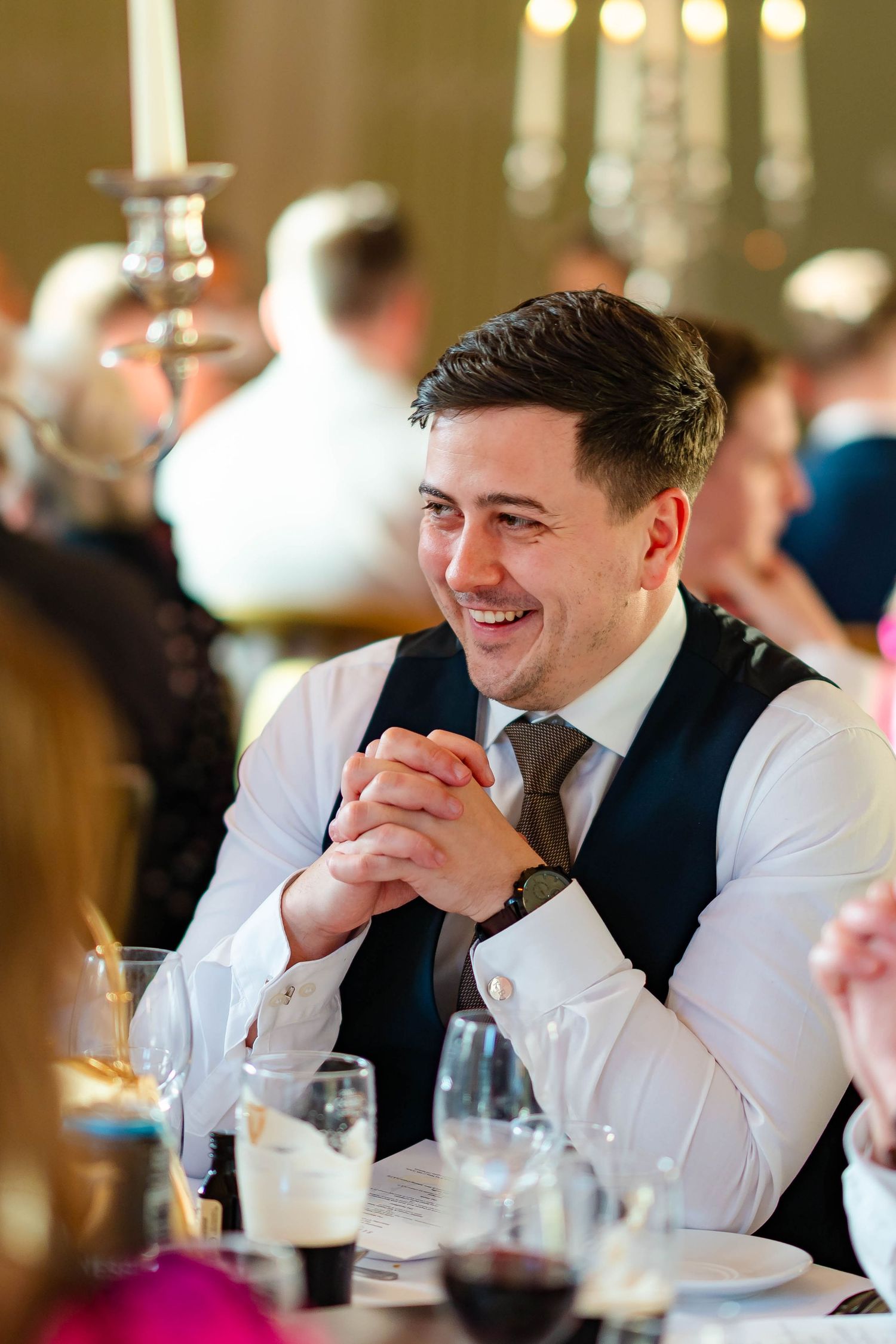 Wedding guest in white shirt and black vest smiles while seated at reception table with wine glasses.