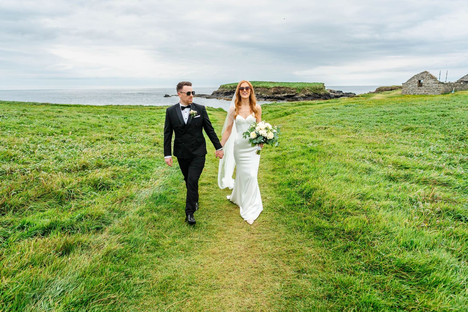 Wedding couple strolls along grassy coastal path with ocean view.