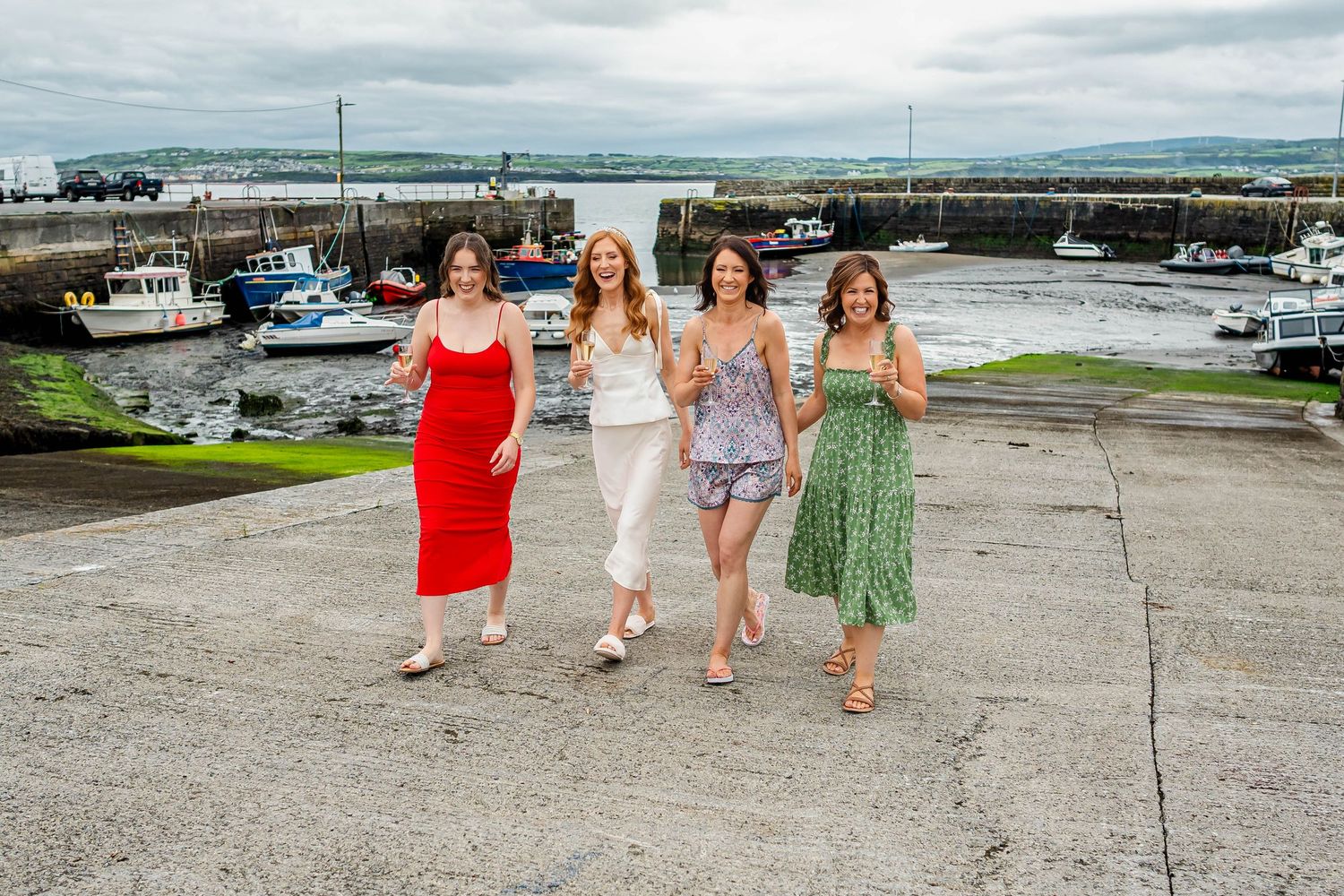 Friends in colorful dresses walk together along a coastal path with boats visible in the harbor.