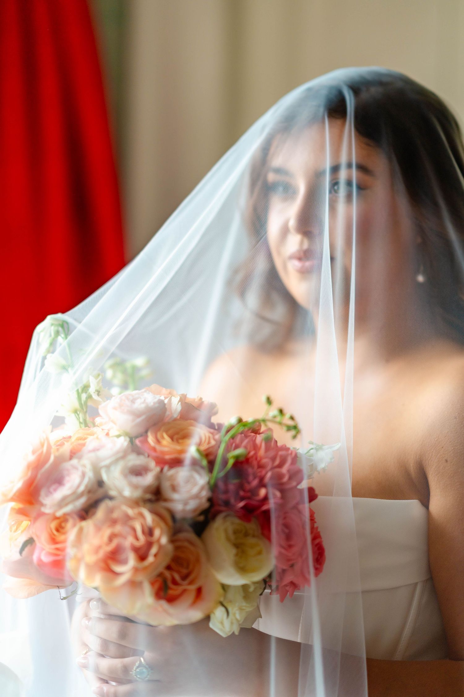 A bride in a white veil holds a beautiful bouquet of peach roses and pink flowers against a red and beige backdrop.