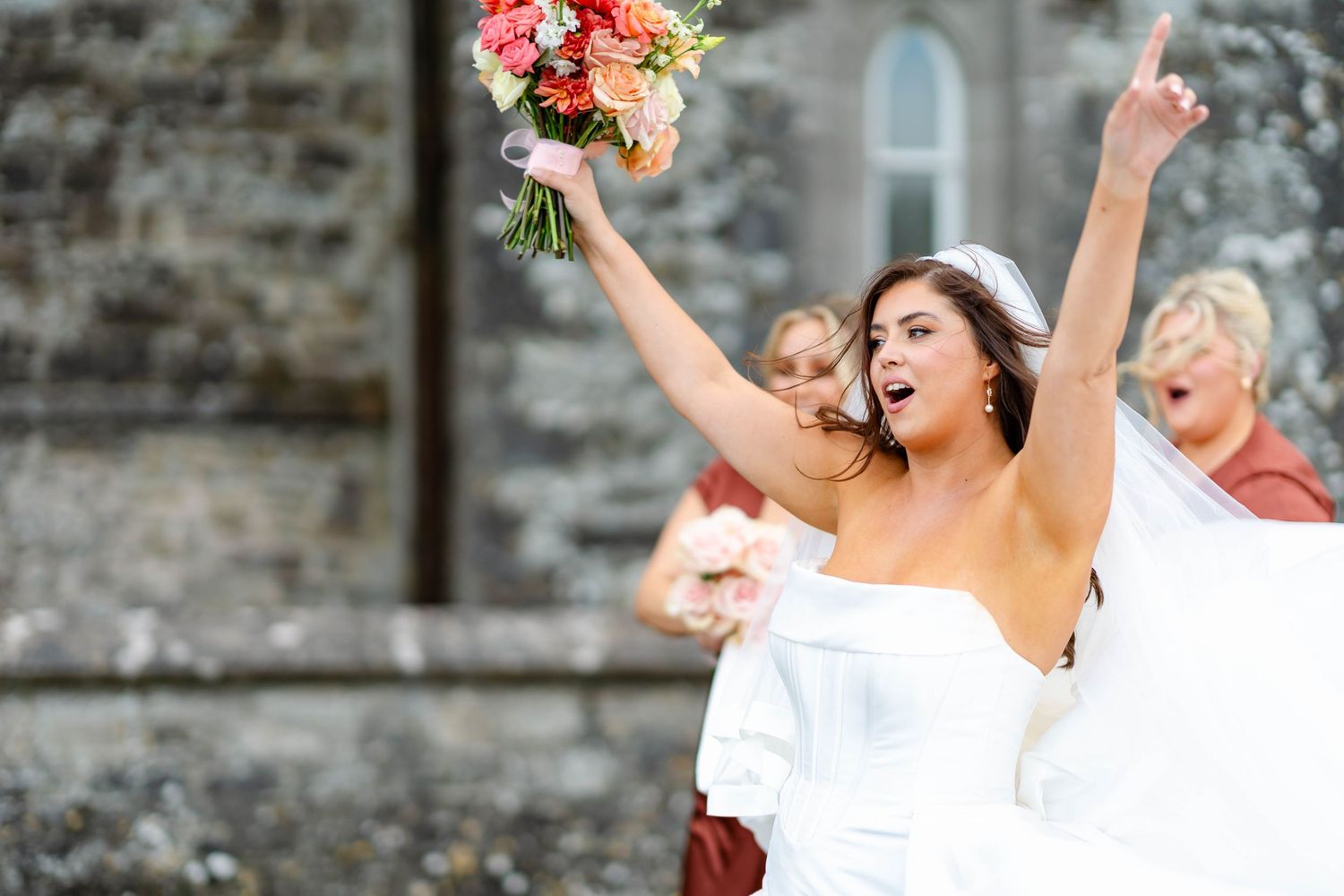 A joyful wedding party celebrates with raised bouquets outside a stone building.