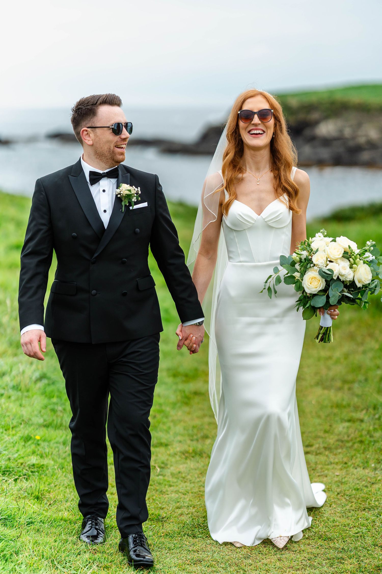 Wedding couple strolls on grassy seaside path wearing black tuxedo and white dress.
