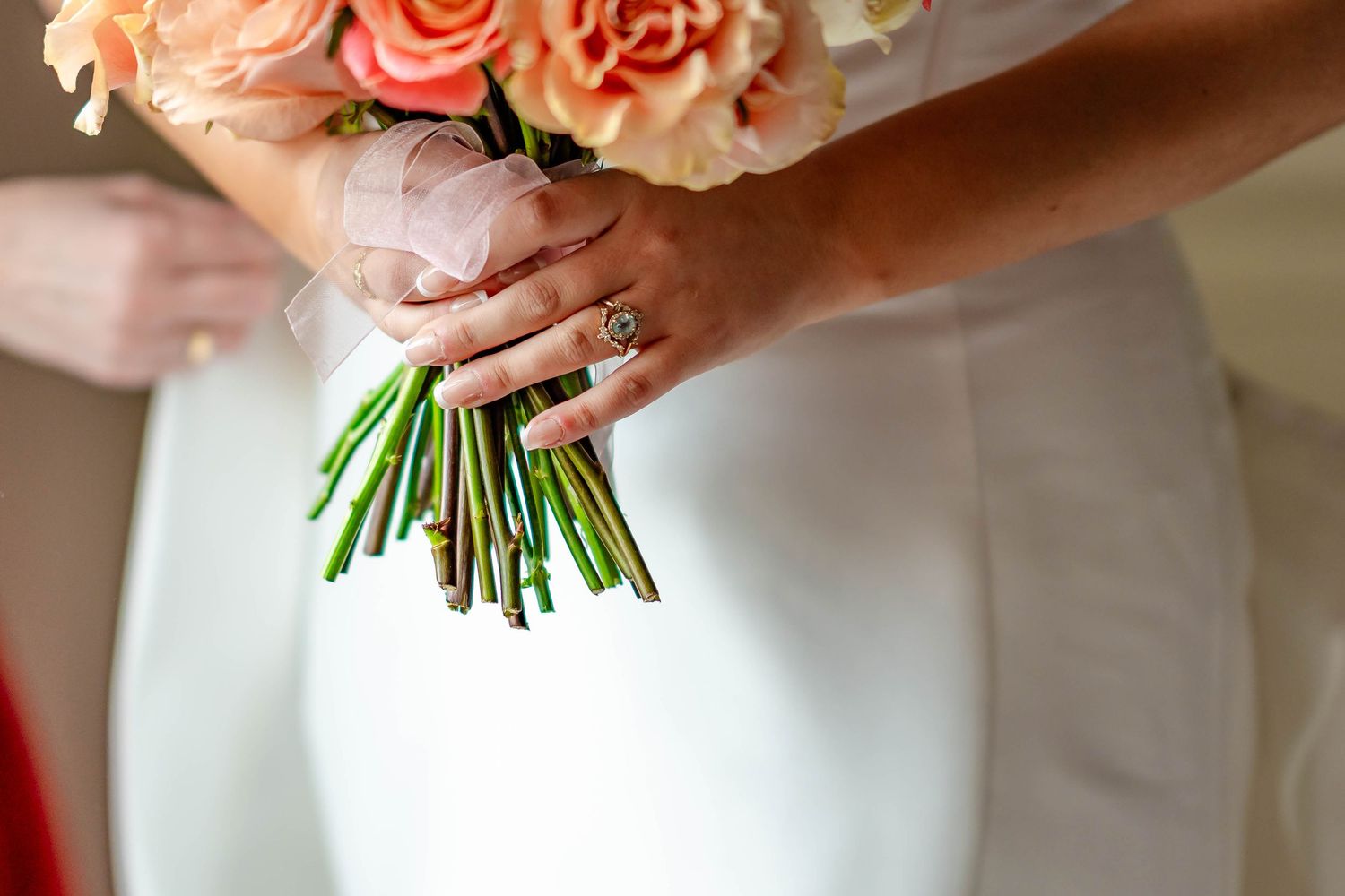 A bridal bouquet of coral and peach roses with white accents being held against a white wedding dress.