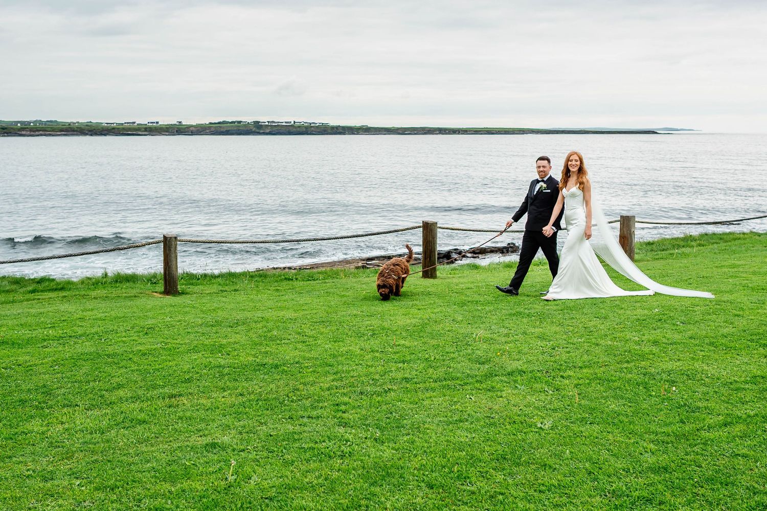 A wedding couple walks along a scenic coastal path with green fields and ocean views.