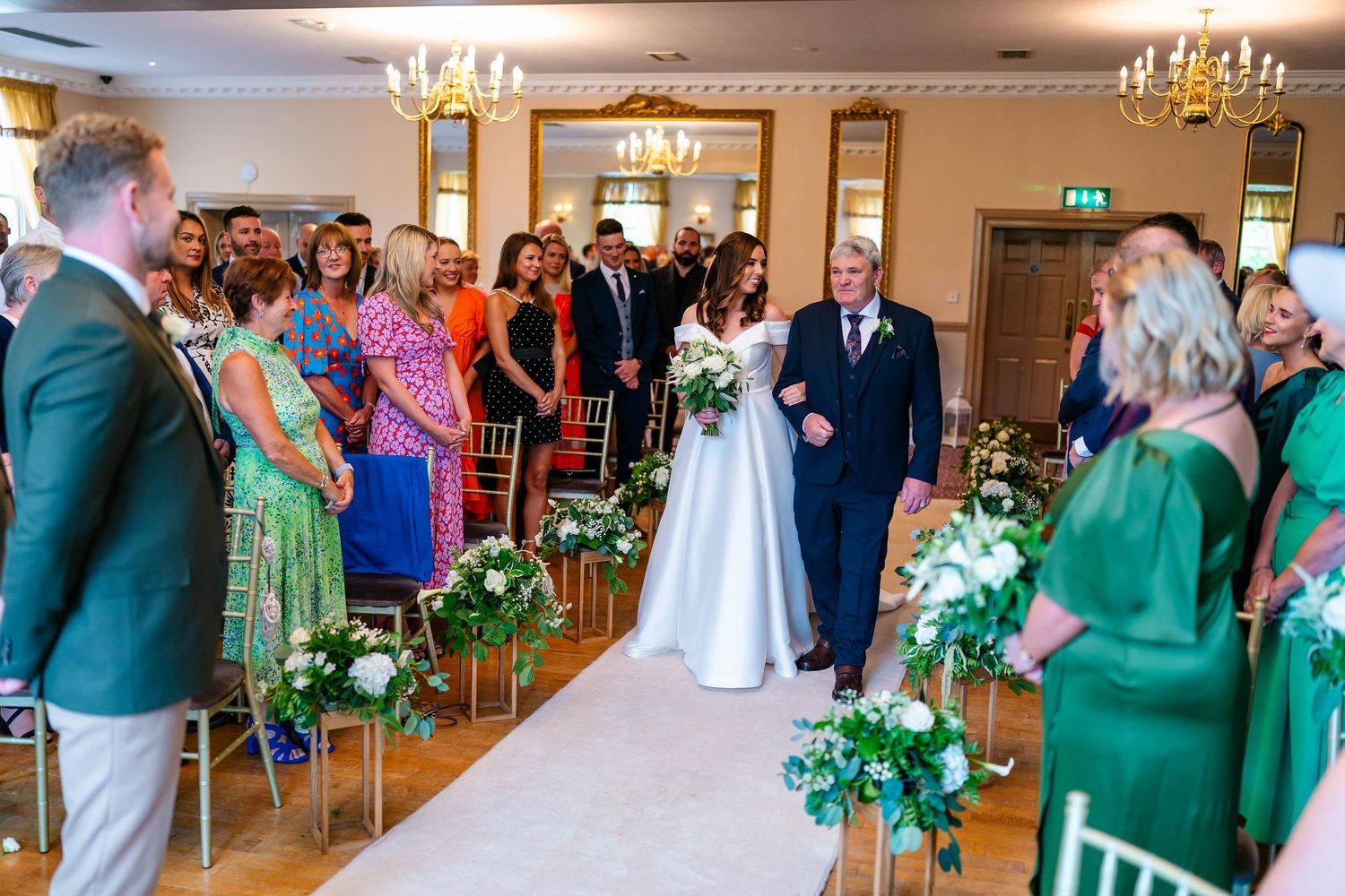 Wedding ceremony in an elegant ballroom with emerald green bridesmaid dresses and white floral decor.