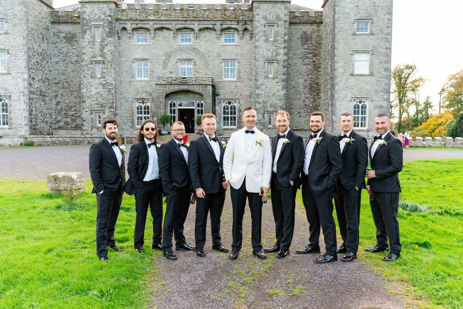 Wedding party in formal attire poses together on the lawn of a historic stone castle or estate.