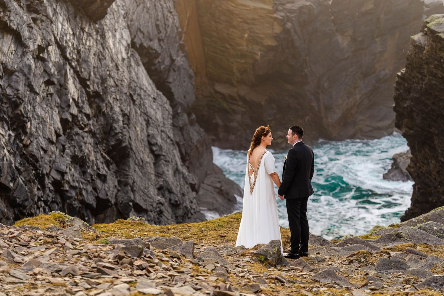 A couple in wedding attire stands on rocky cliffs overlooking dramatic ocean waves at sunset.