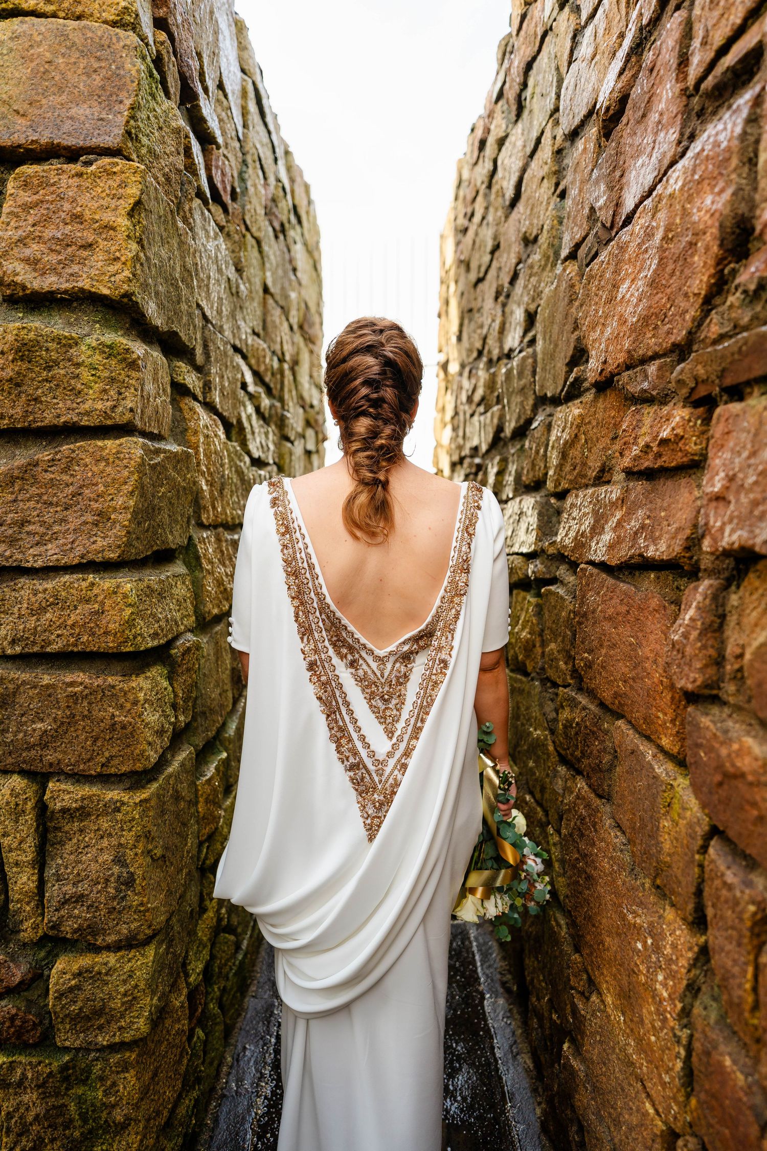 Back view of wedding dress with intricate beaded detail between rustic stone walls.