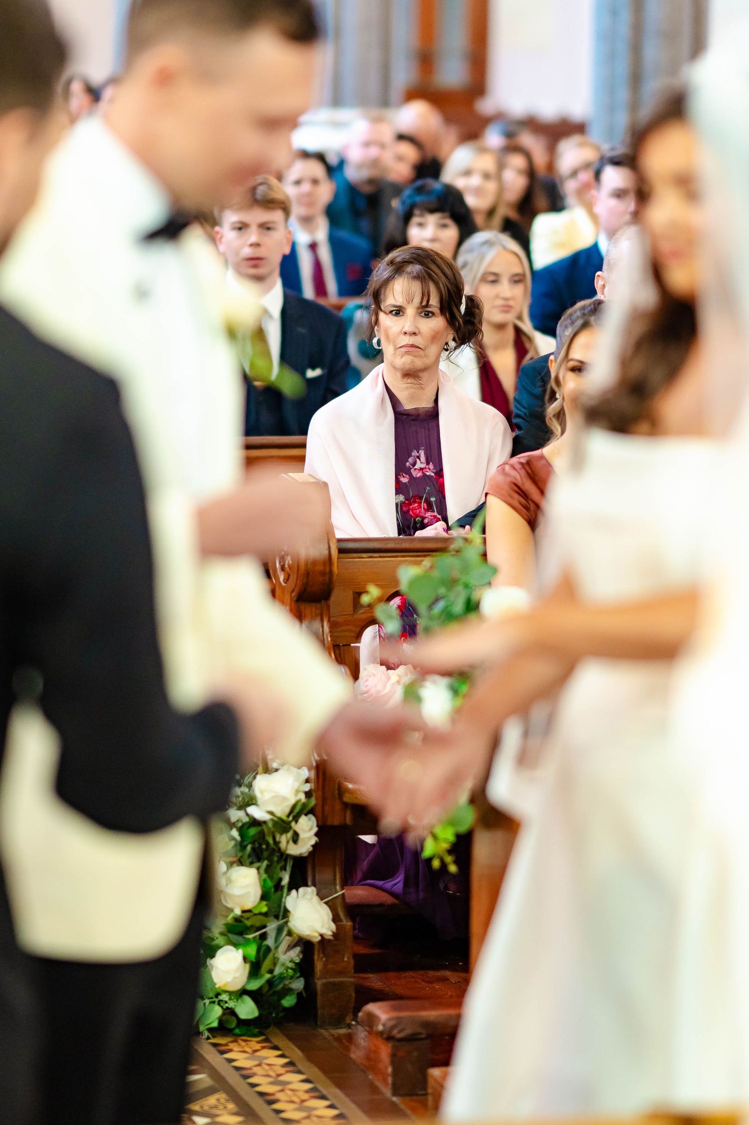 A wedding ceremony in a church with guests seated in wooden pews and floral decorations along the aisle.