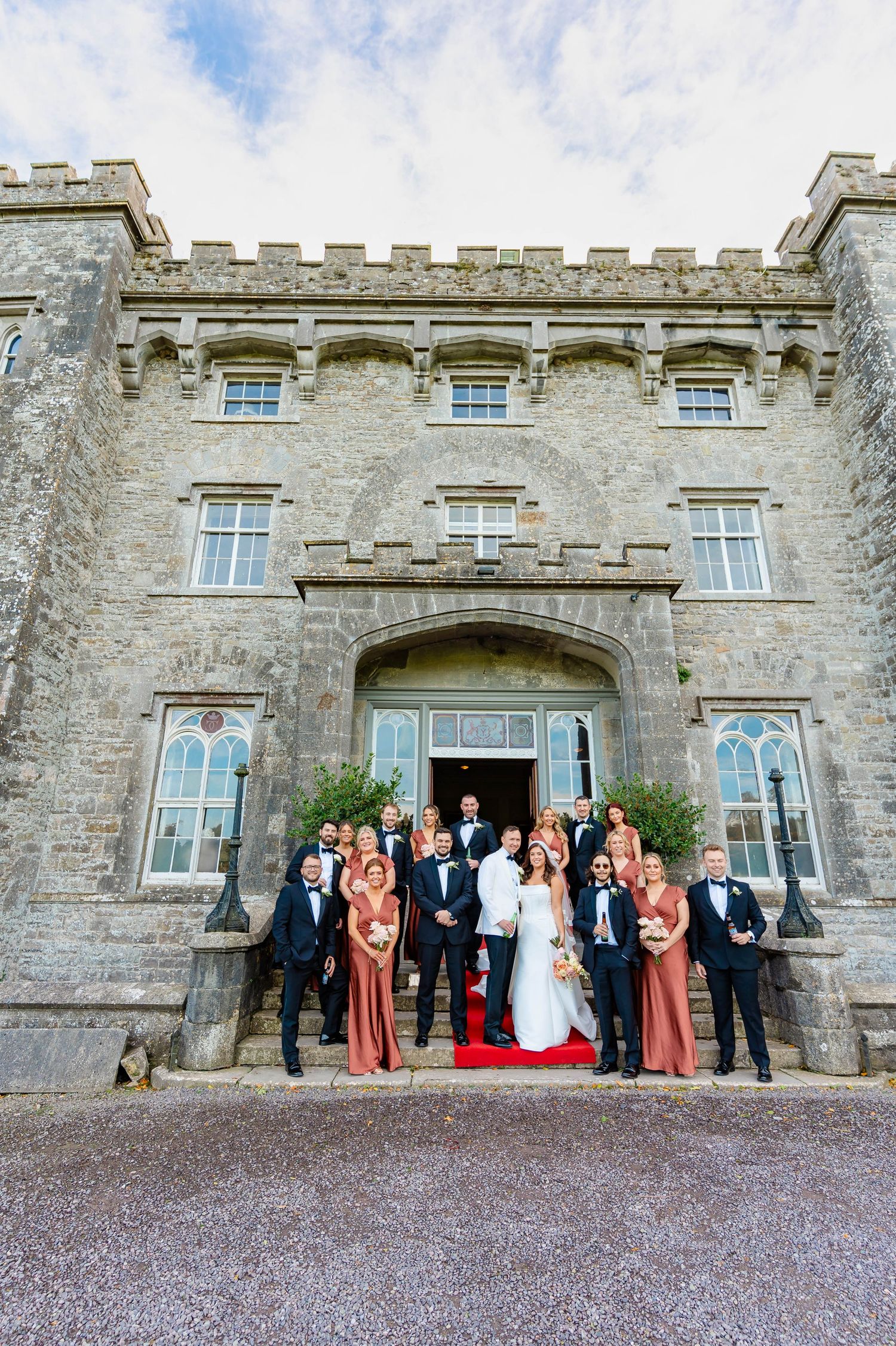 Wedding party poses outside historic stone castle with gray walls and windows in formal attire.