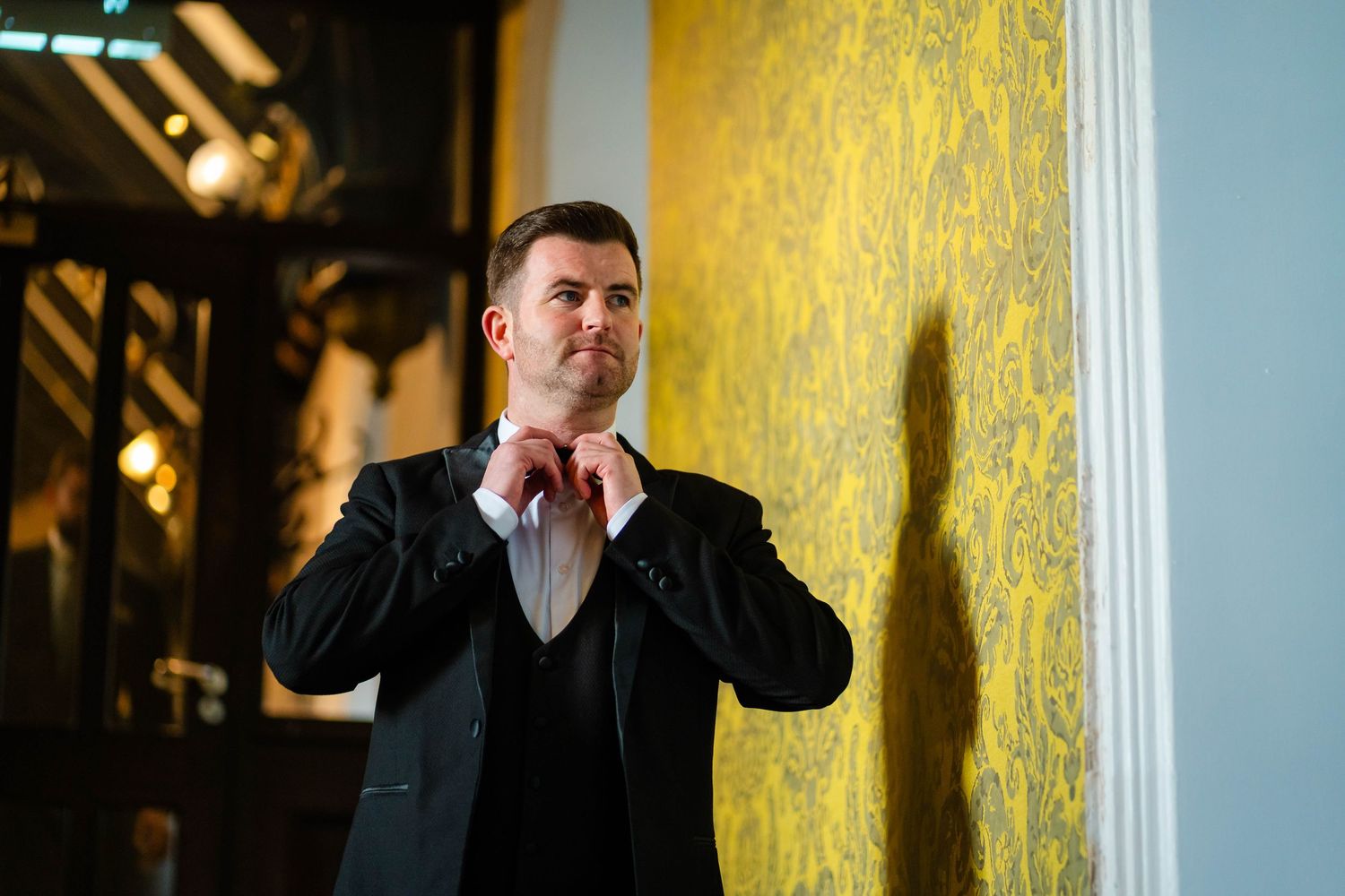 Person in formal wear posing against a yellow patterned wallpaper background.