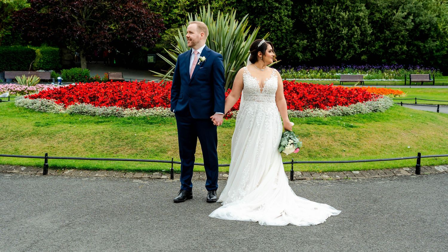 Panoramic view of newlyweds walking in a park with red flower beds and green landscaping.