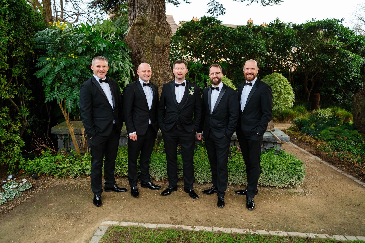 Five groomsmen in black tuxedos pose on garden path.