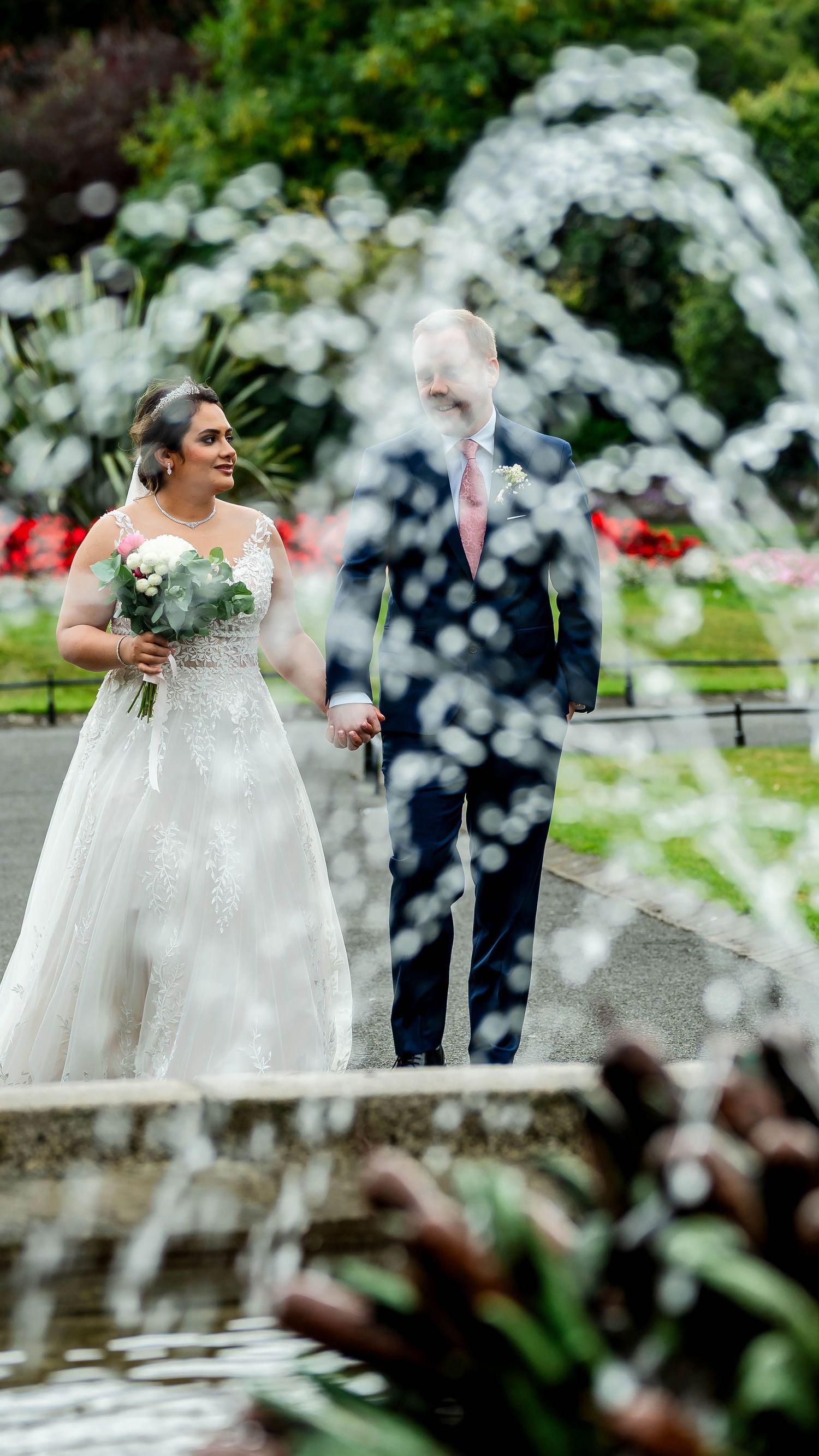 A couple stands near a fountain with water spray creating a dreamy effect in a garden setting.