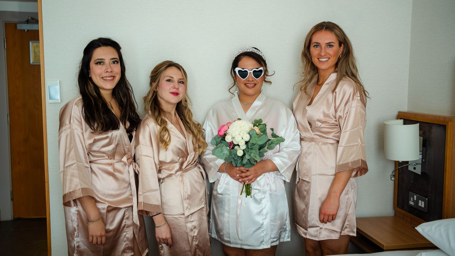 Wedding party members in matching pink satin robes gathered in a bright room.