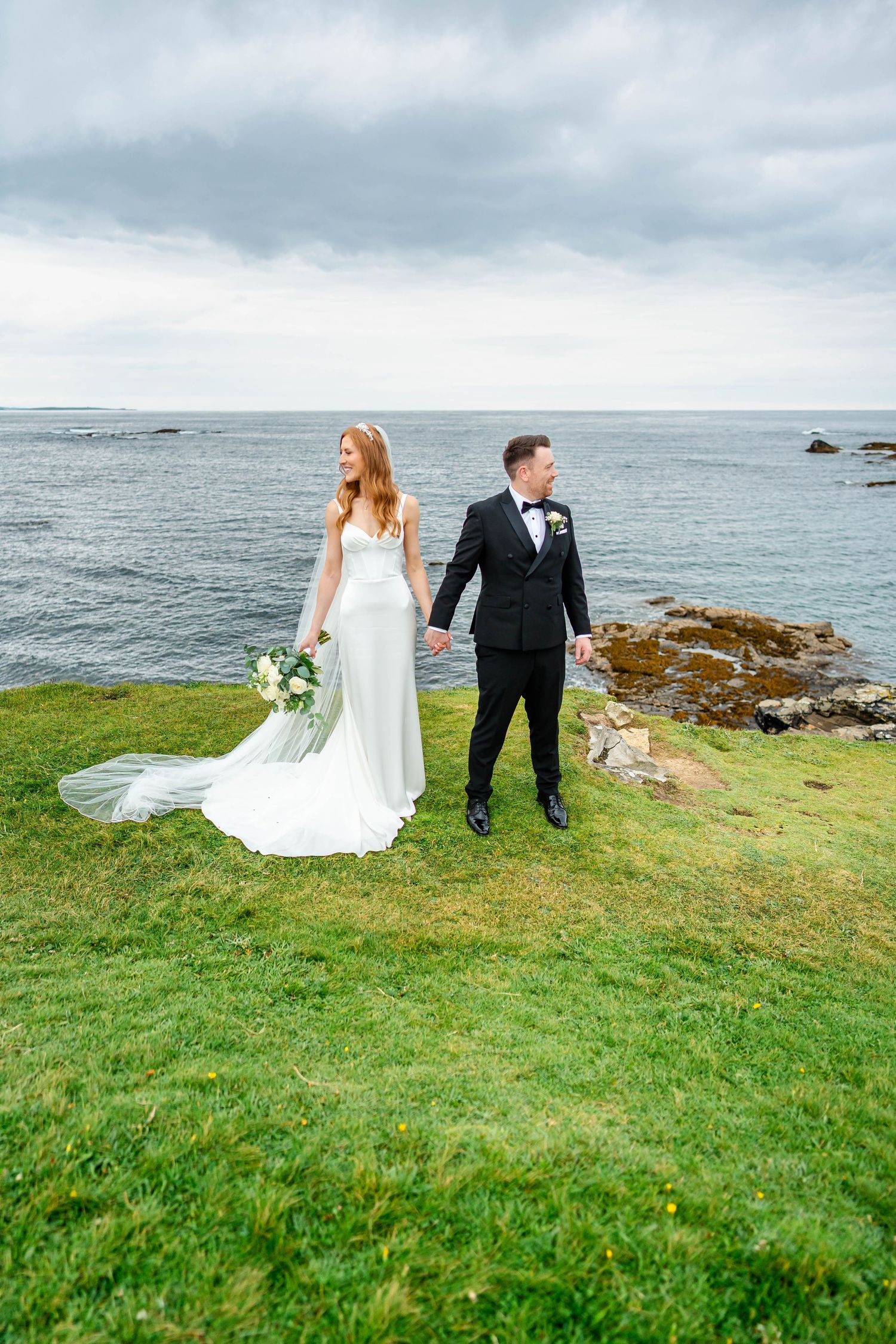 Wedding couple stands on dramatic coastal cliffs overlooking the ocean.