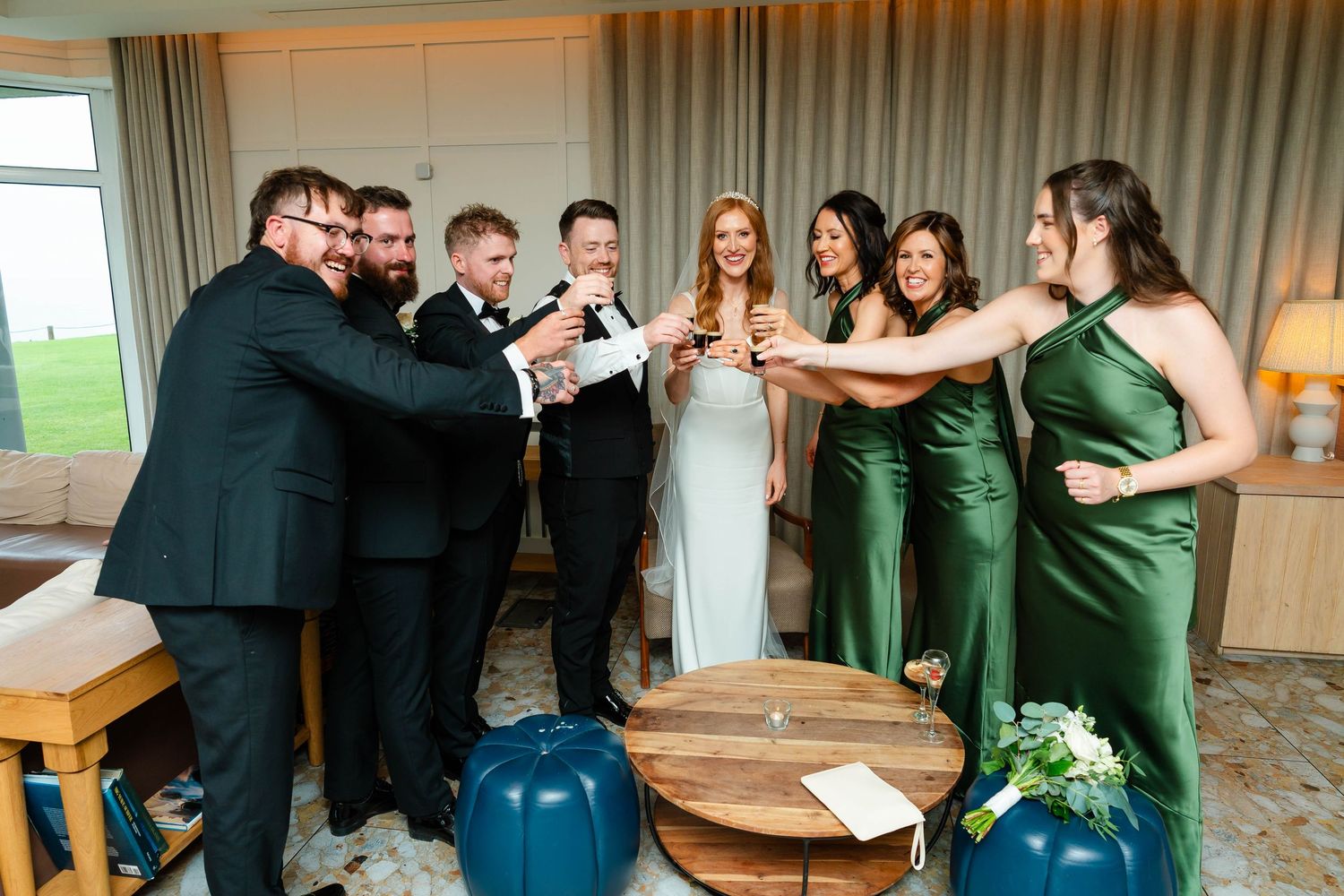 Wedding party toasts together in a modern venue with emerald green bridesmaid dresses.