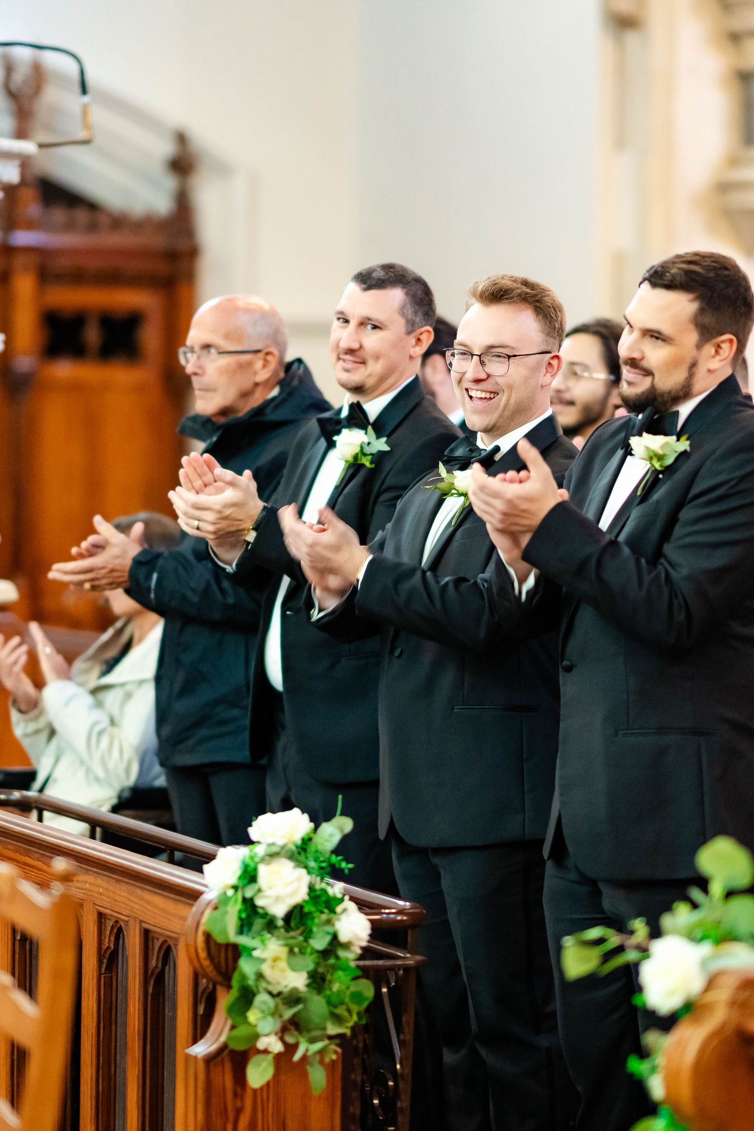 Groomsmen in black tuxedos stand in church pews during wedding ceremony with white and green floral decorations.