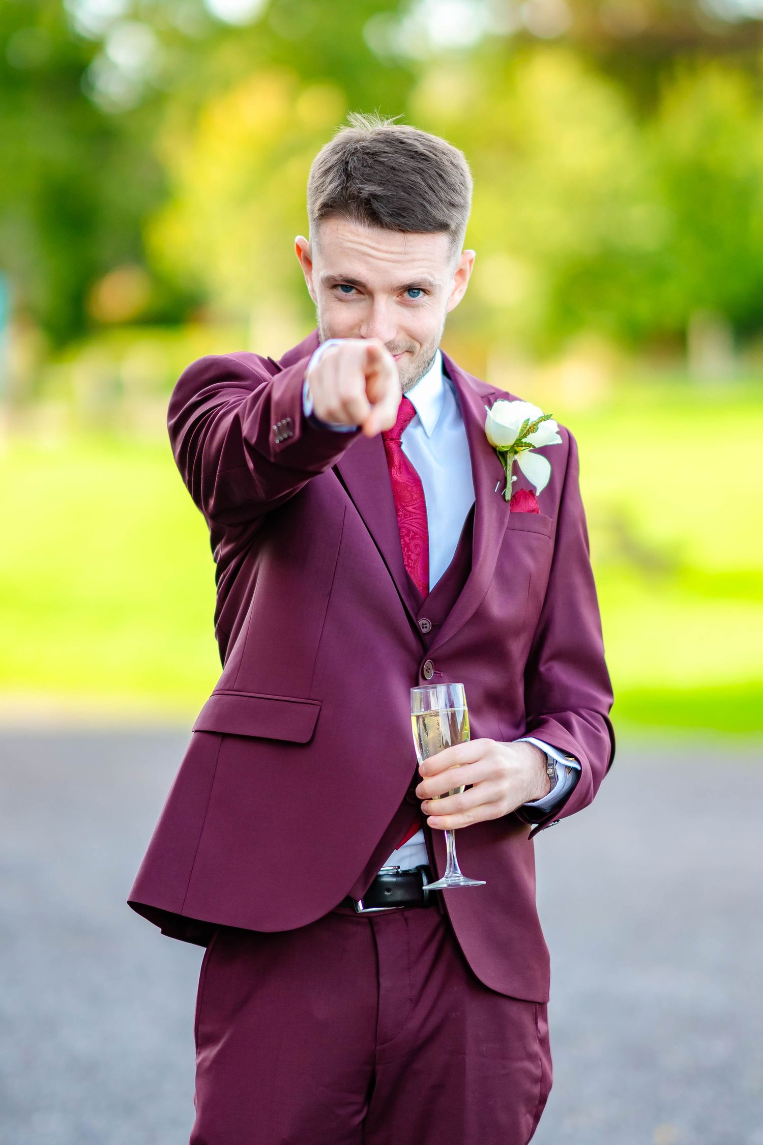 A person in a burgundy suit and blue tie holds a champagne glass while making celebratory gestures outdoors.