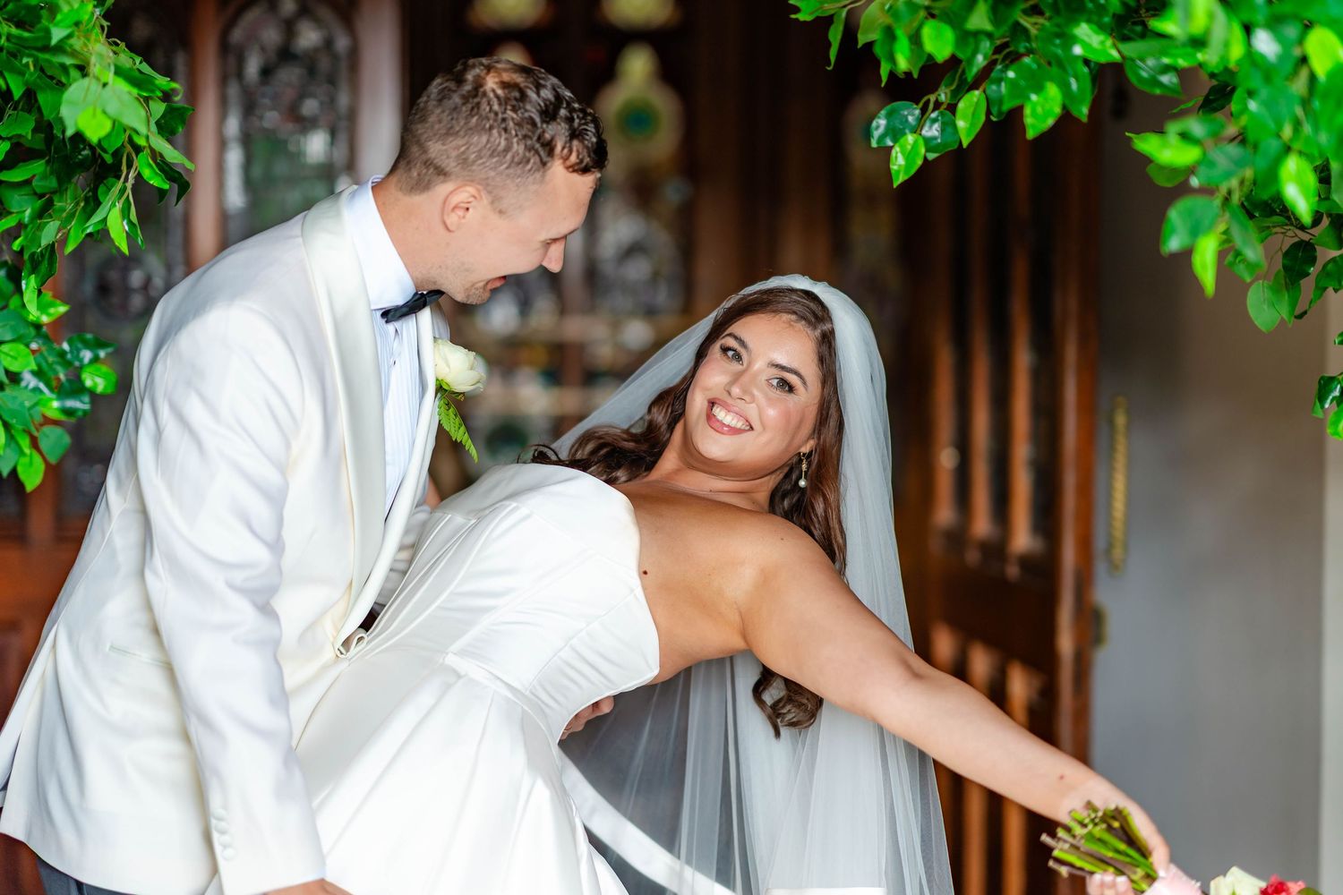 A romantic wedding couple shares intimate moments under green foliage at a rustic wooden venue.