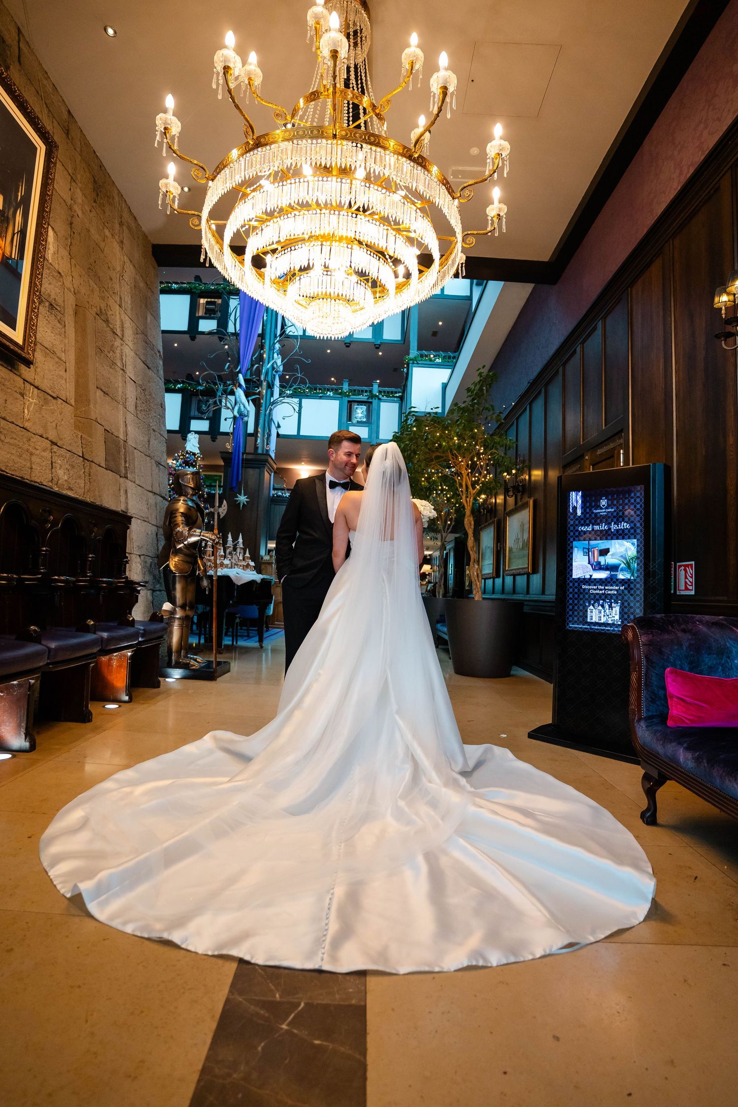 Bride in flowing white gown stands under elegant crystal chandelier in grand venue.