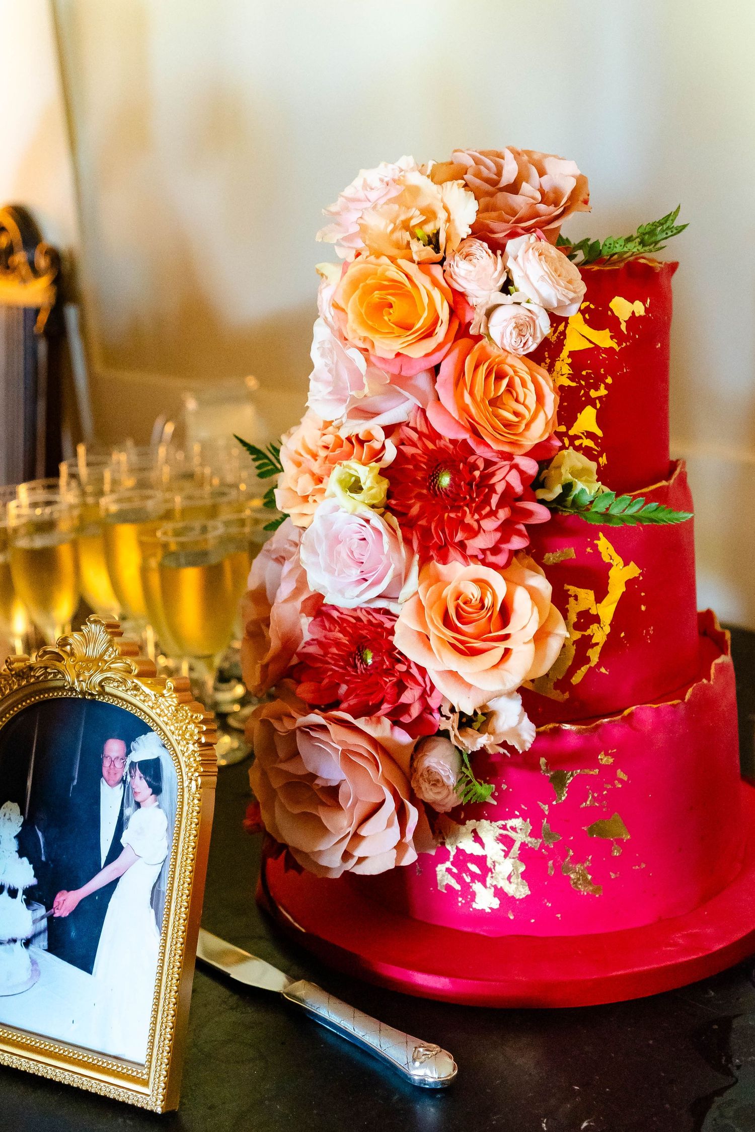 Red wedding cake decorated with gold accents and peach roses sits beside a framed photograph and wine glasses.