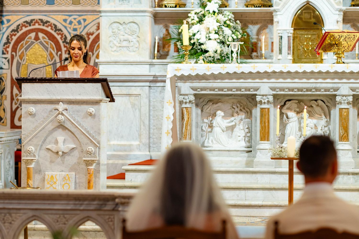 People attend a religious service in a church with ornate marble altar and religious artwork decorations.
