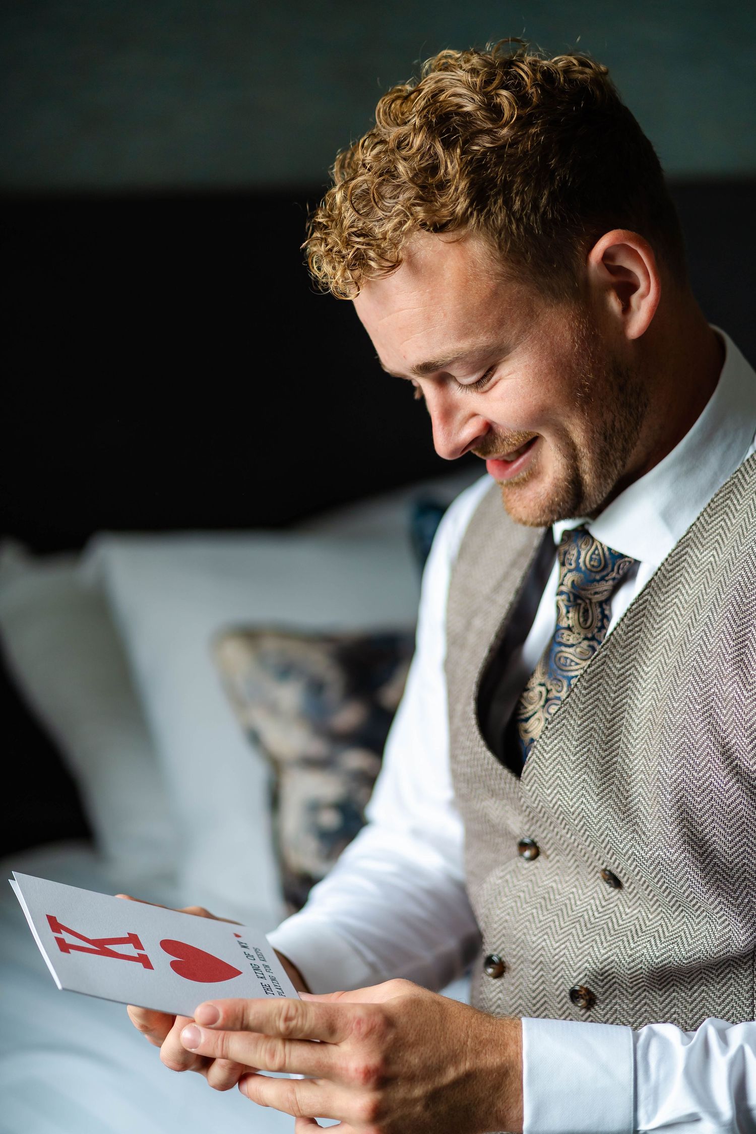 Groom in grey vest smiles while reading a playing card message.