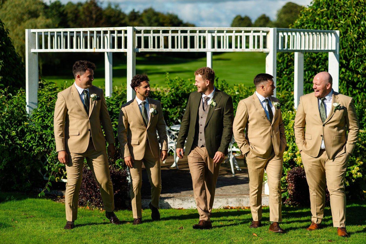 Wedding party in tan suits poses together on lush green lawn.