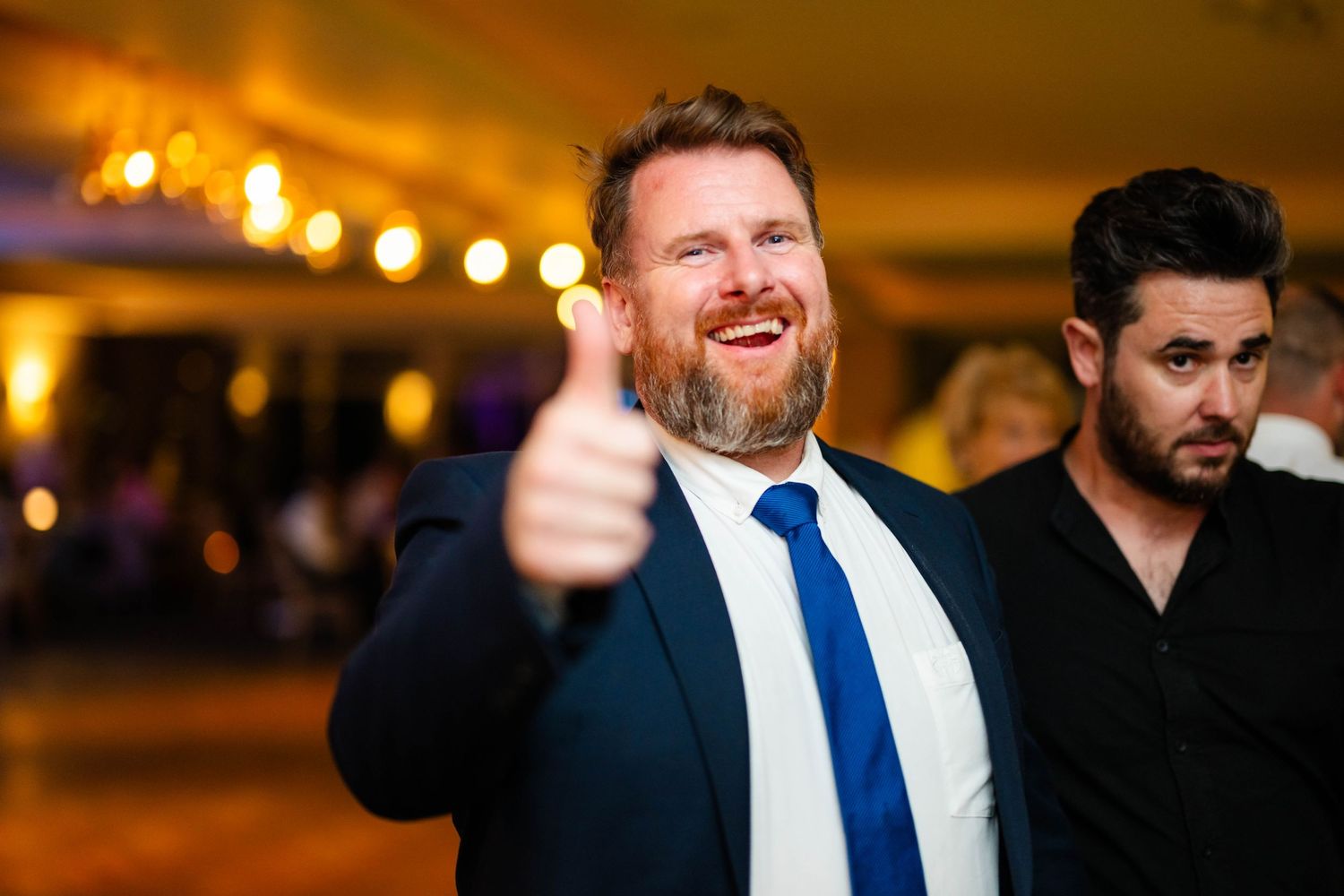 Wedding guest in navy suit and blue tie gives enthusiastic thumbs up at reception celebration.