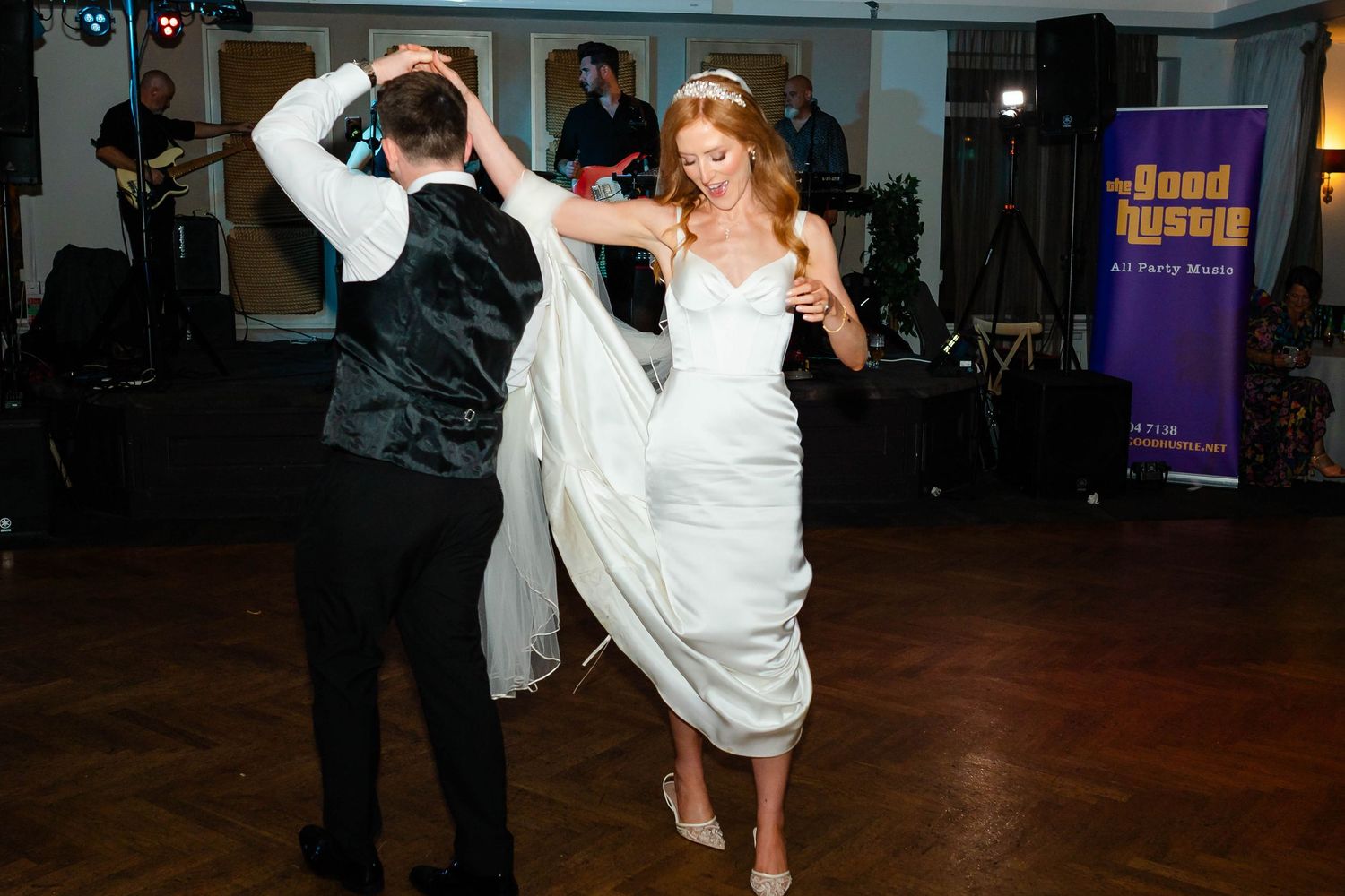 Couple dancing energetically at a wedding reception with colorful lighting and band in the background.
