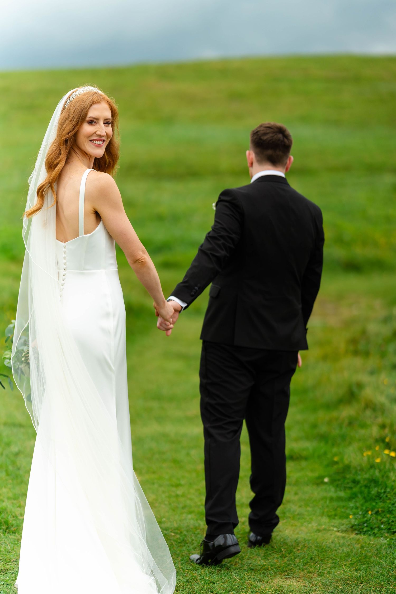Candid wedding photography showing couple walking through green coastal meadow.