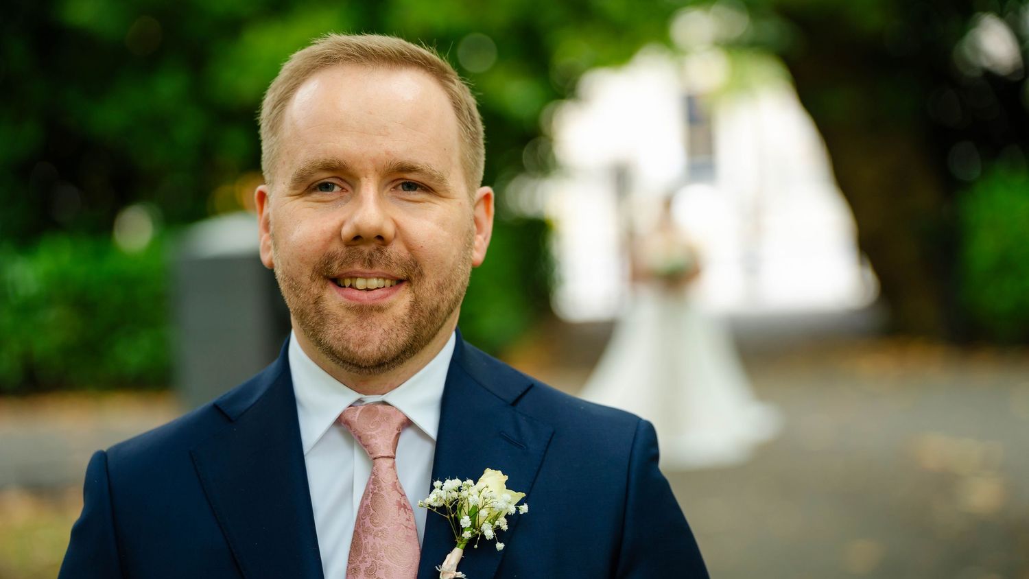 Groom in navy suit with pink tie and boutonniere stands in outdoor setting.