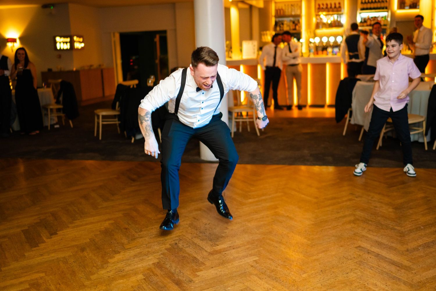 People performing coordinated dance moves on a wooden dance floor at a wedding venue.