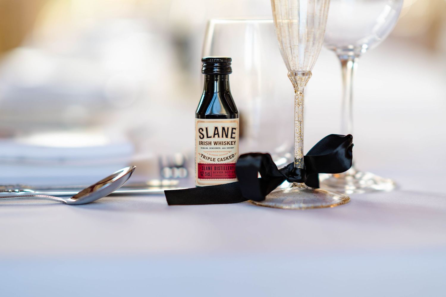 A bottle of Slane Irish Whiskey sits on a white tablecloth next to empty champagne glasses and dining utensils.
