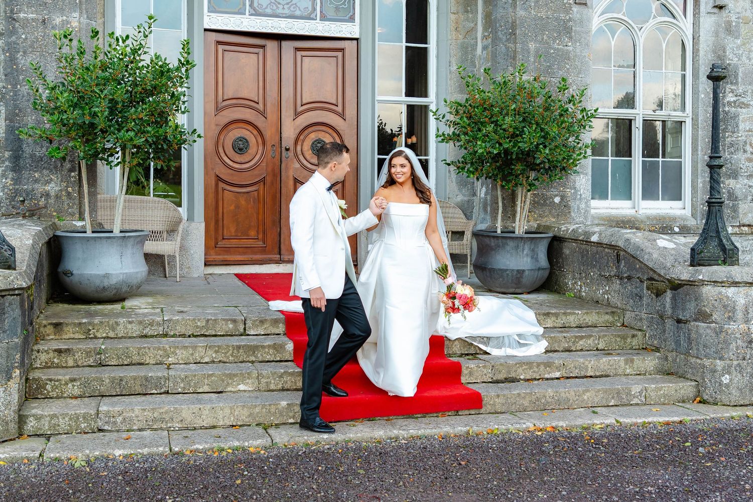 A couple in wedding attire poses on red-carpeted stone steps in front of an elegant wooden door with potted plants.