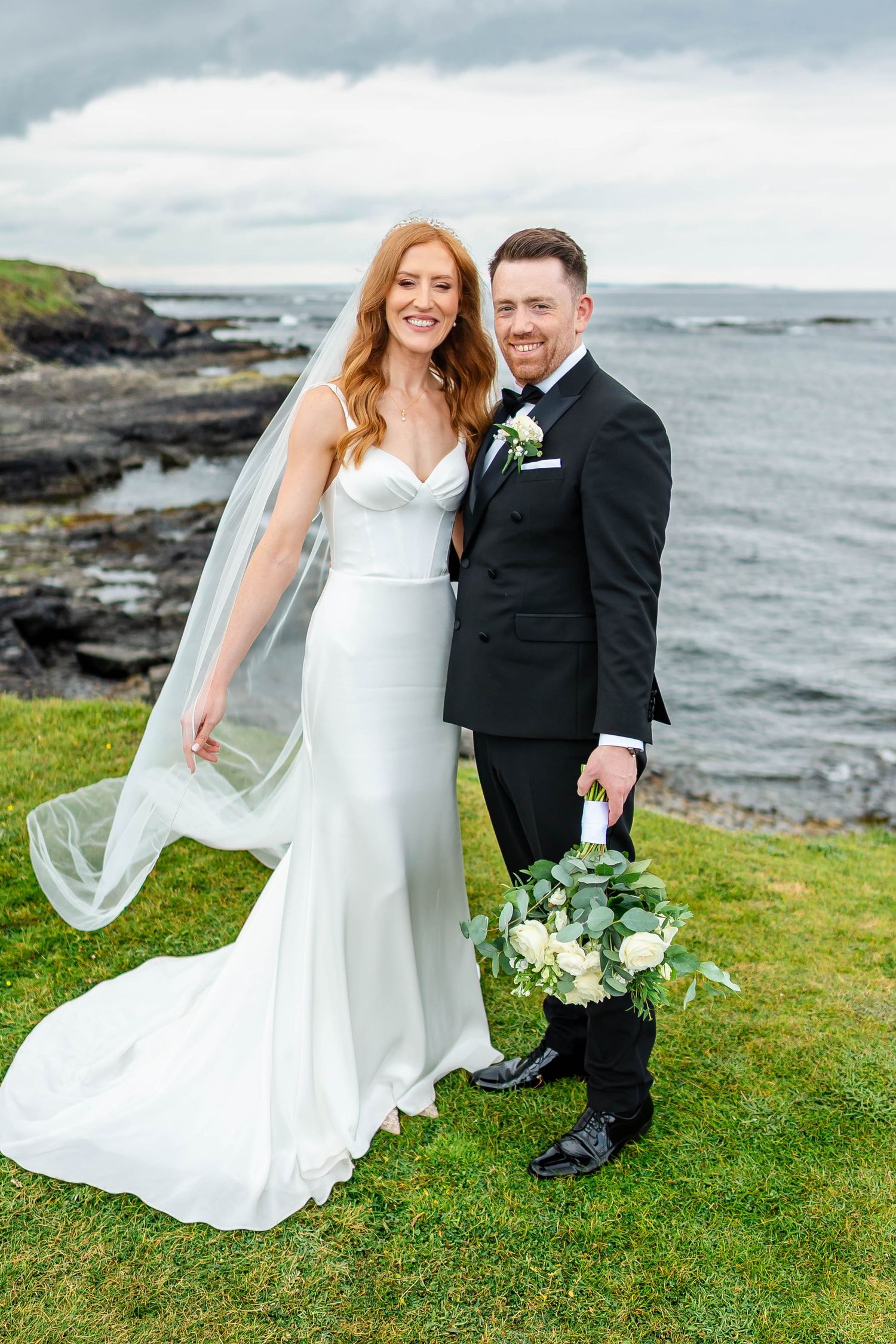 Coastal wedding portrait with flowing white gown and lush bouquet against ocean backdrop.