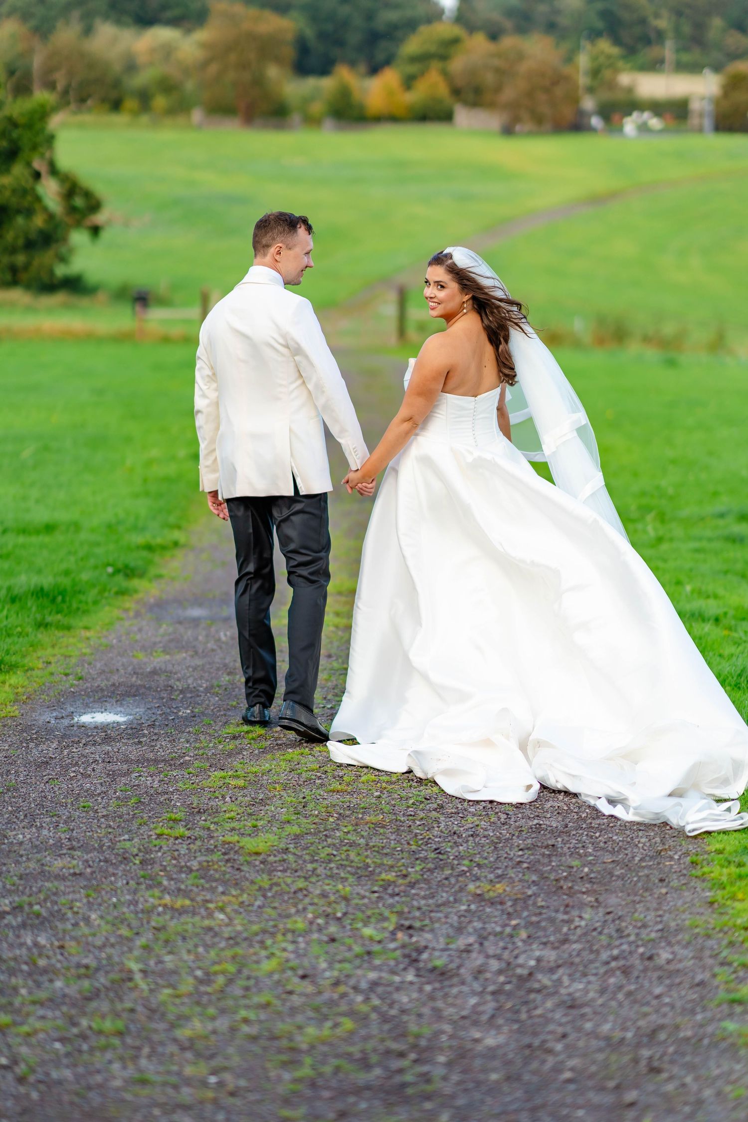 Bride and groom walking along a path through green fields while her white wedding dress billows in the wind.