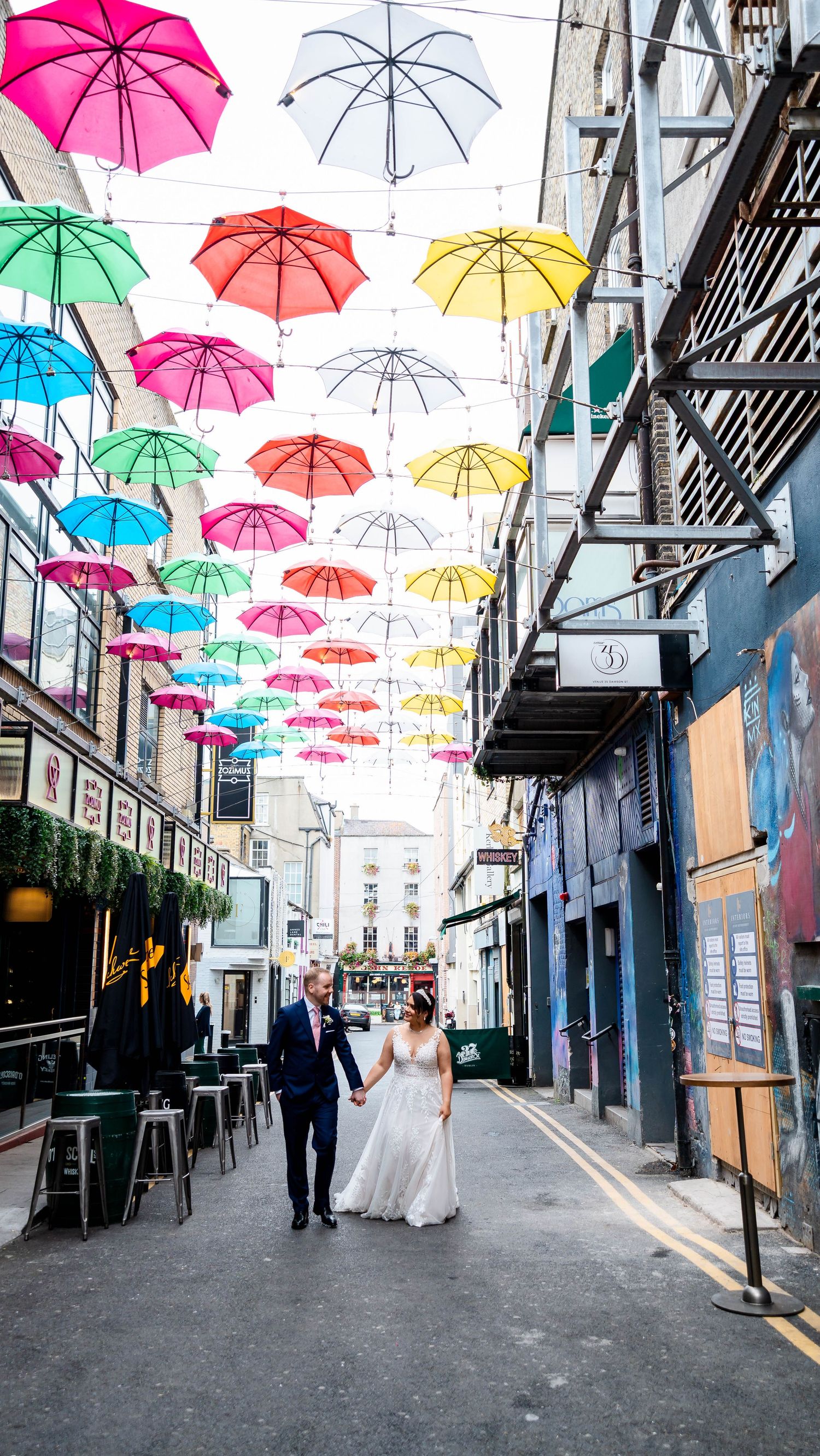 Urban street art installation featuring pink, blue, yellow and white umbrellas hanging above a narrow alley.